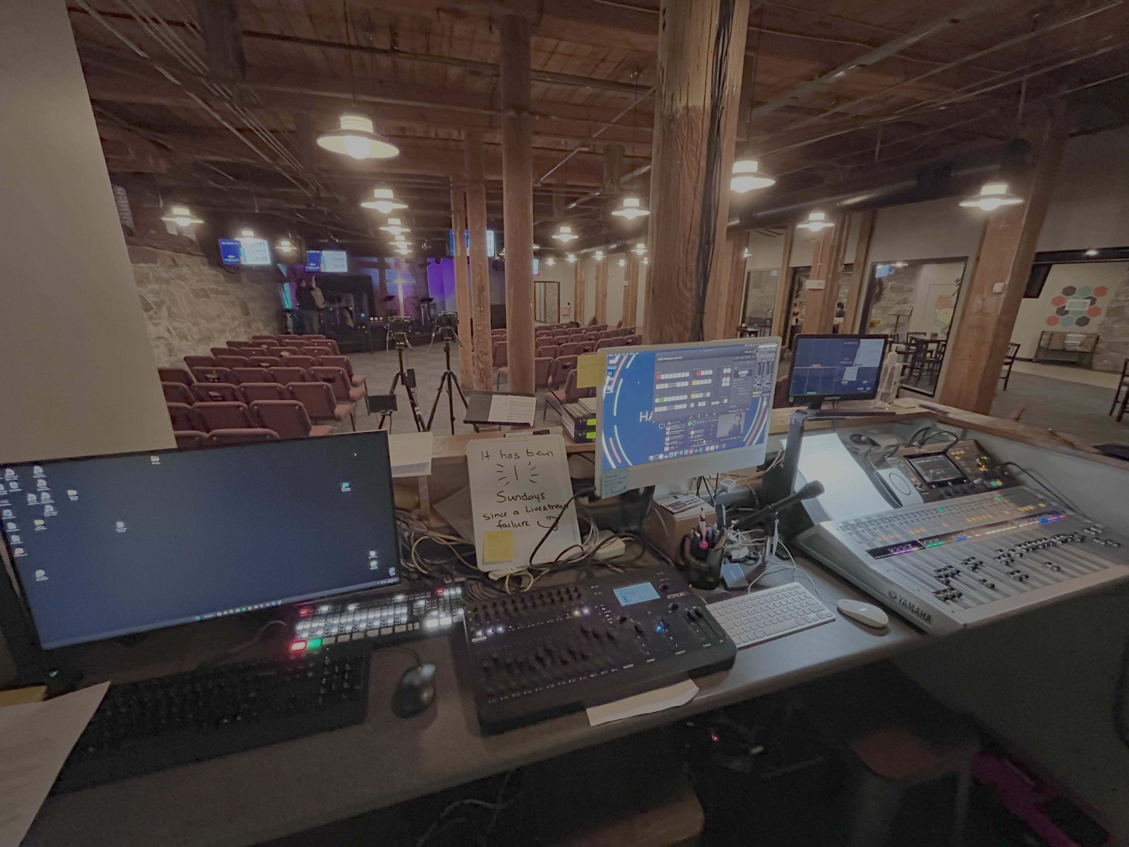 The image shows a control booth equipped with multiple computer monitors, a soundboard, and keyboards overlooking a spacious auditorium with seating arranged for an event.