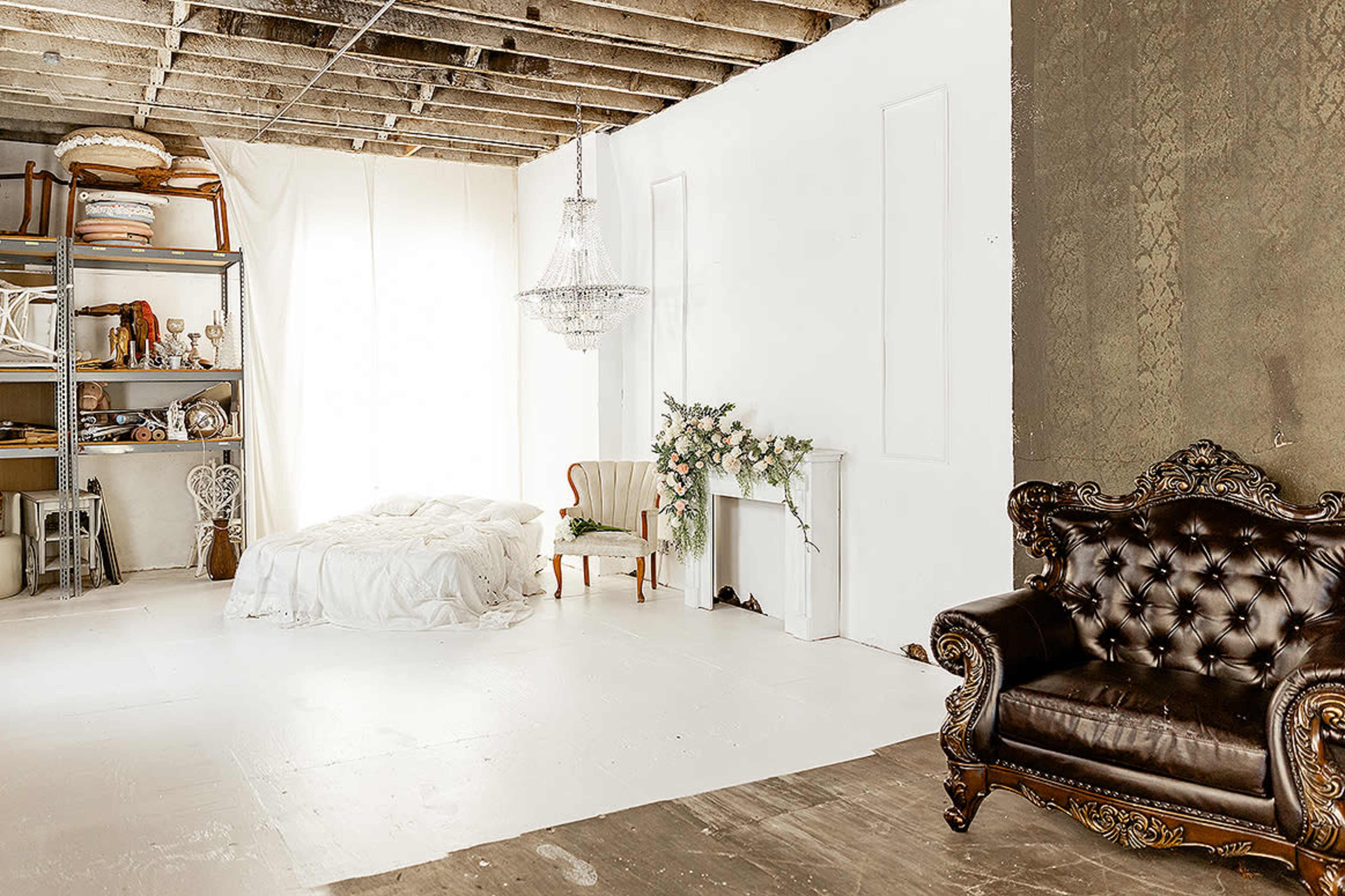 The scene features a spacious, minimally decorated room with a bed, a vintage chair, a chandelier, and a dark upholstered couch against a backdrop of white walls and exposed ceiling beams.