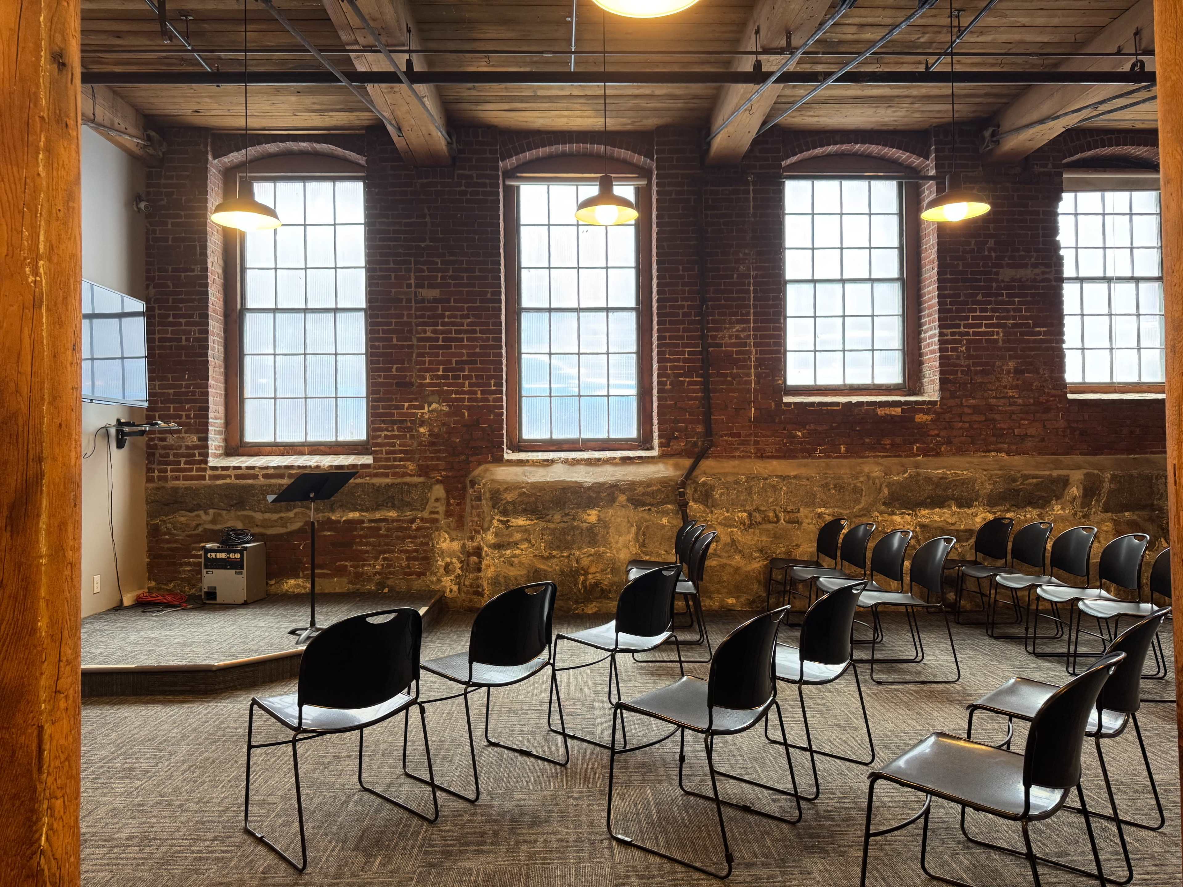 The image shows a room with exposed brick walls, large windows, and a set of black chairs arranged in rows facing a small stage area.