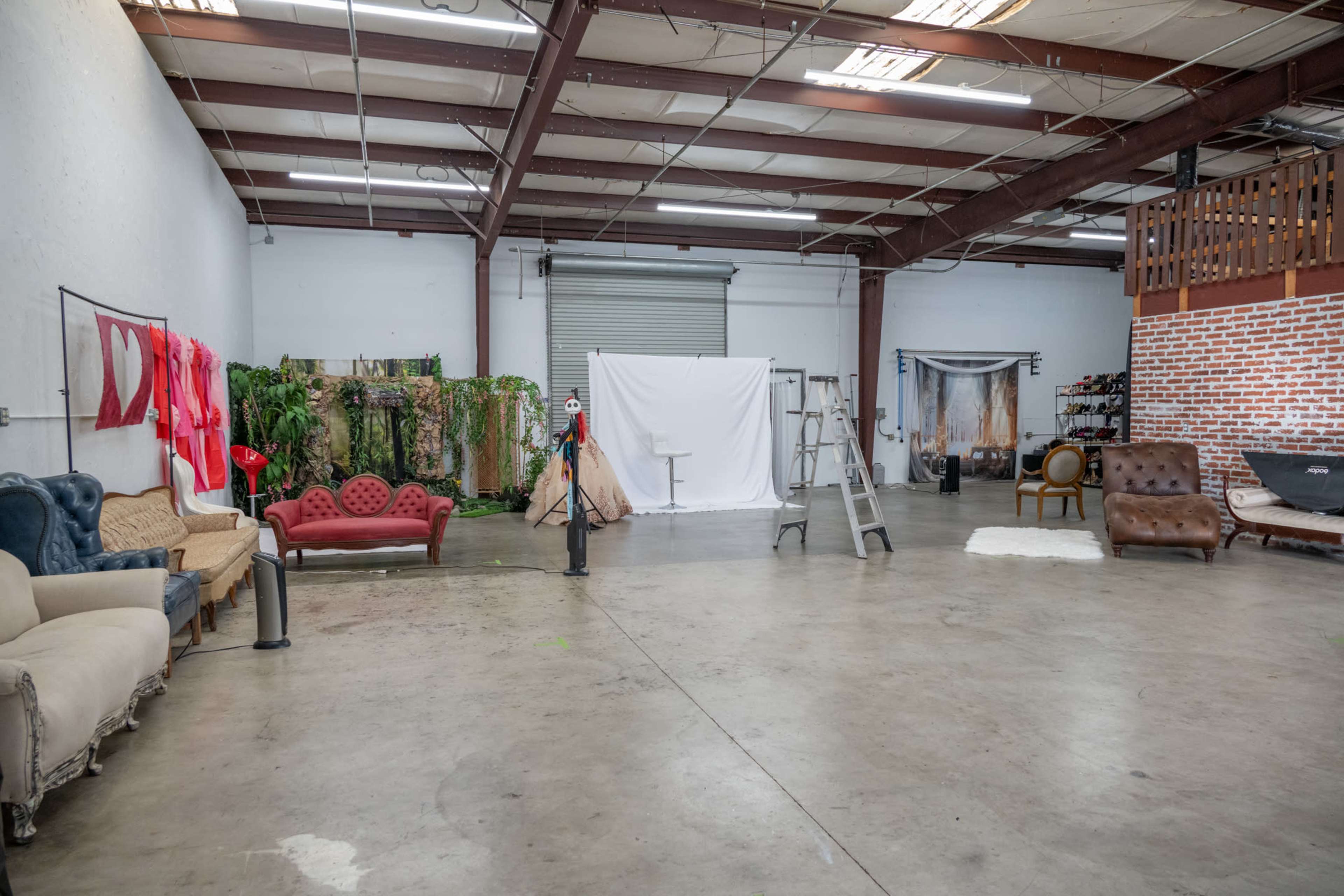 The image shows an empty studio space with various props, including decorative furniture and a white backdrop, set against exposed metal beams and a brick wall.