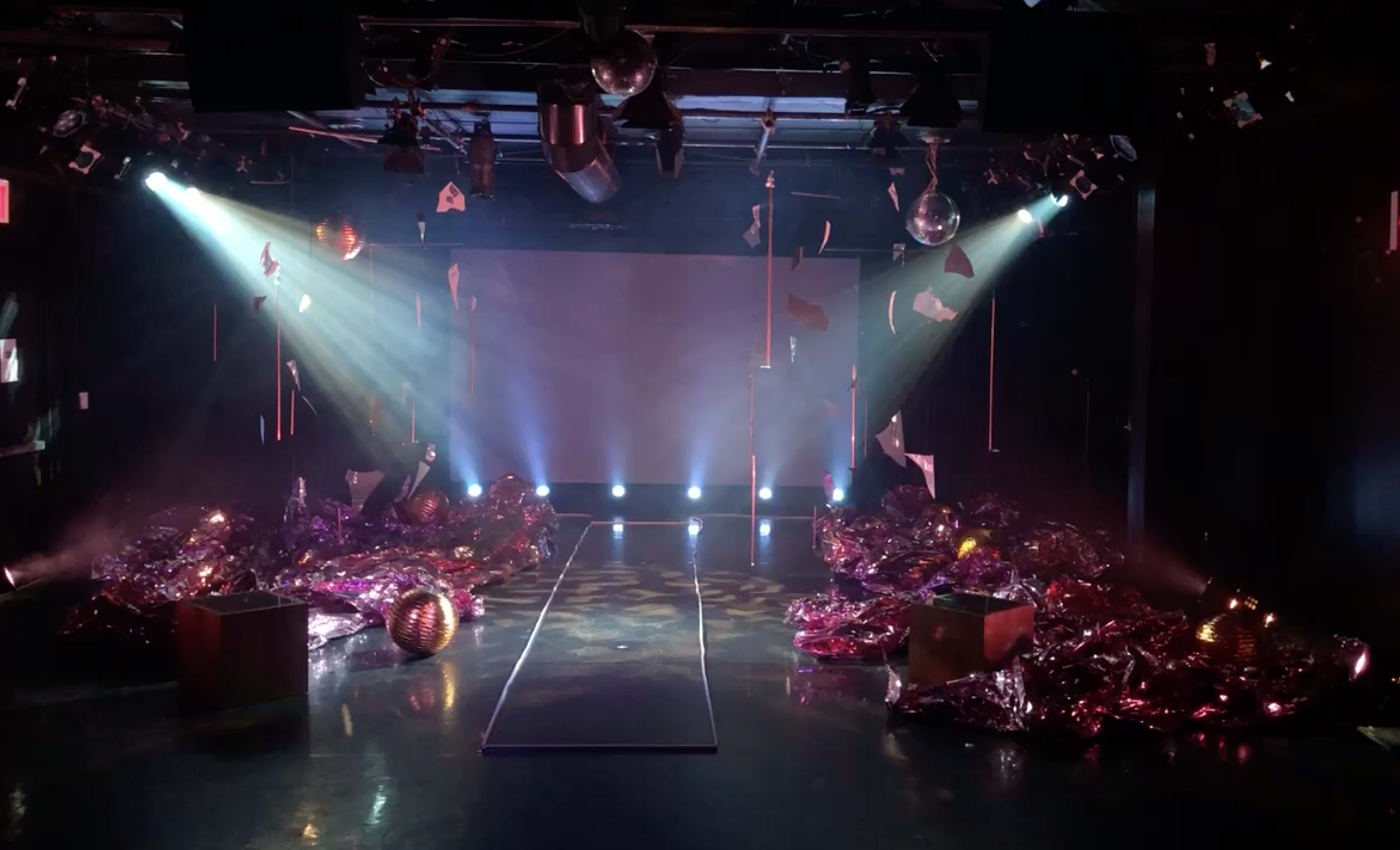 A stage with a central runway surrounded by colorful debris, illuminated by beams of light and disco balls hanging overhead.