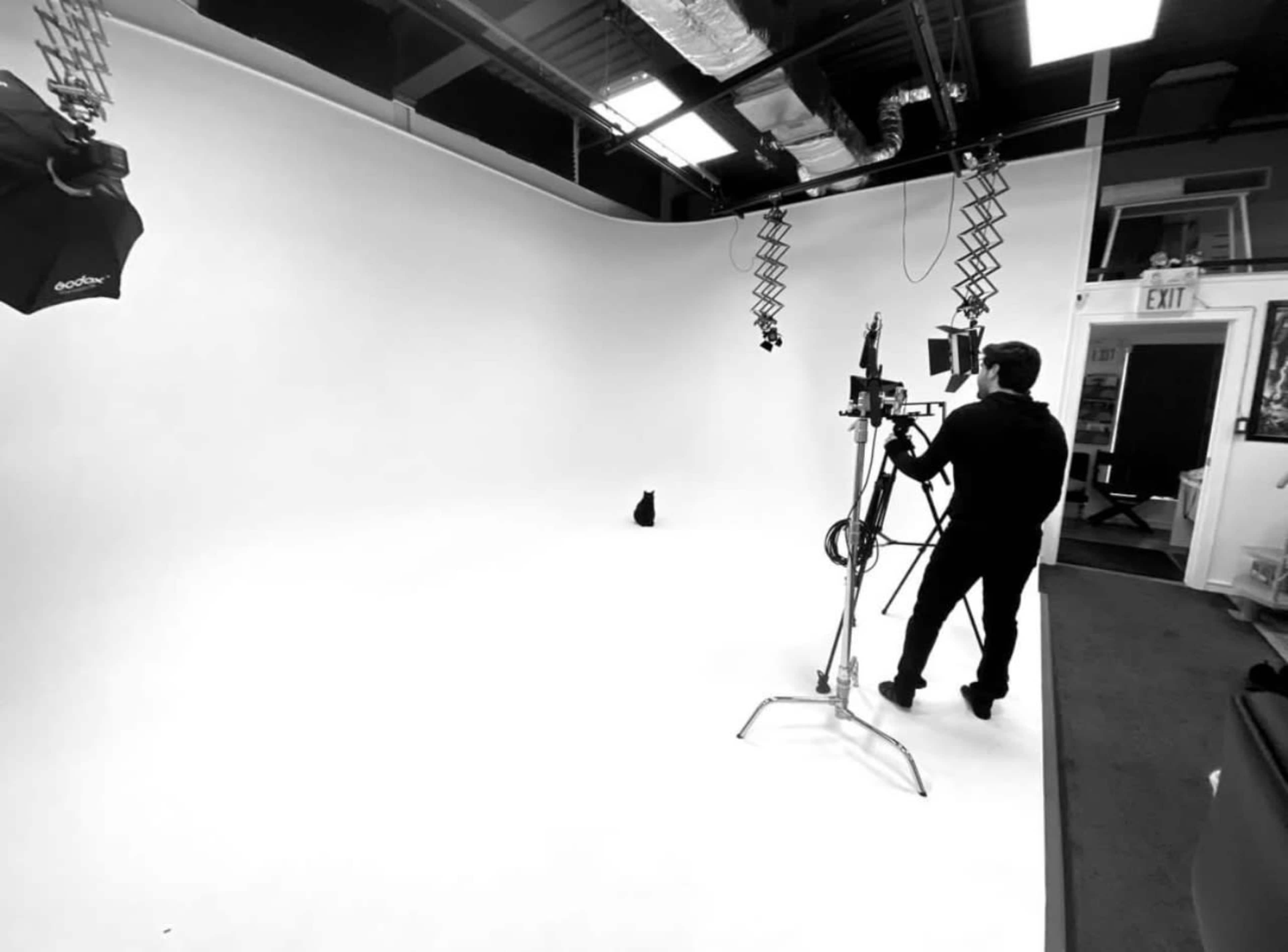 A person operates a camera in a spacious, well-lit studio with a white backdrop and equipment setup for a shoot.