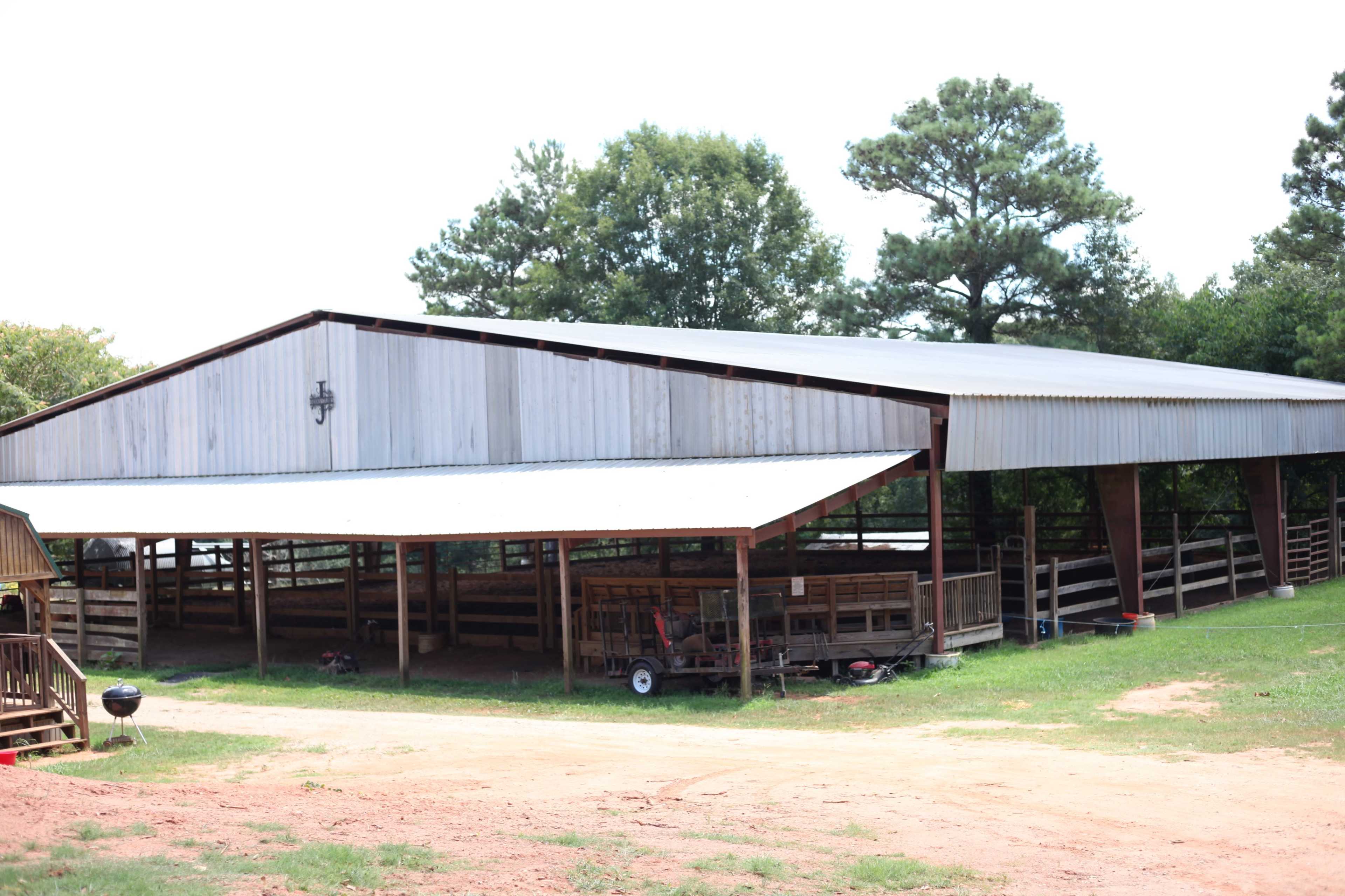 A large metal barn with a peaked roof sits on a grassy area, surrounded by trees.