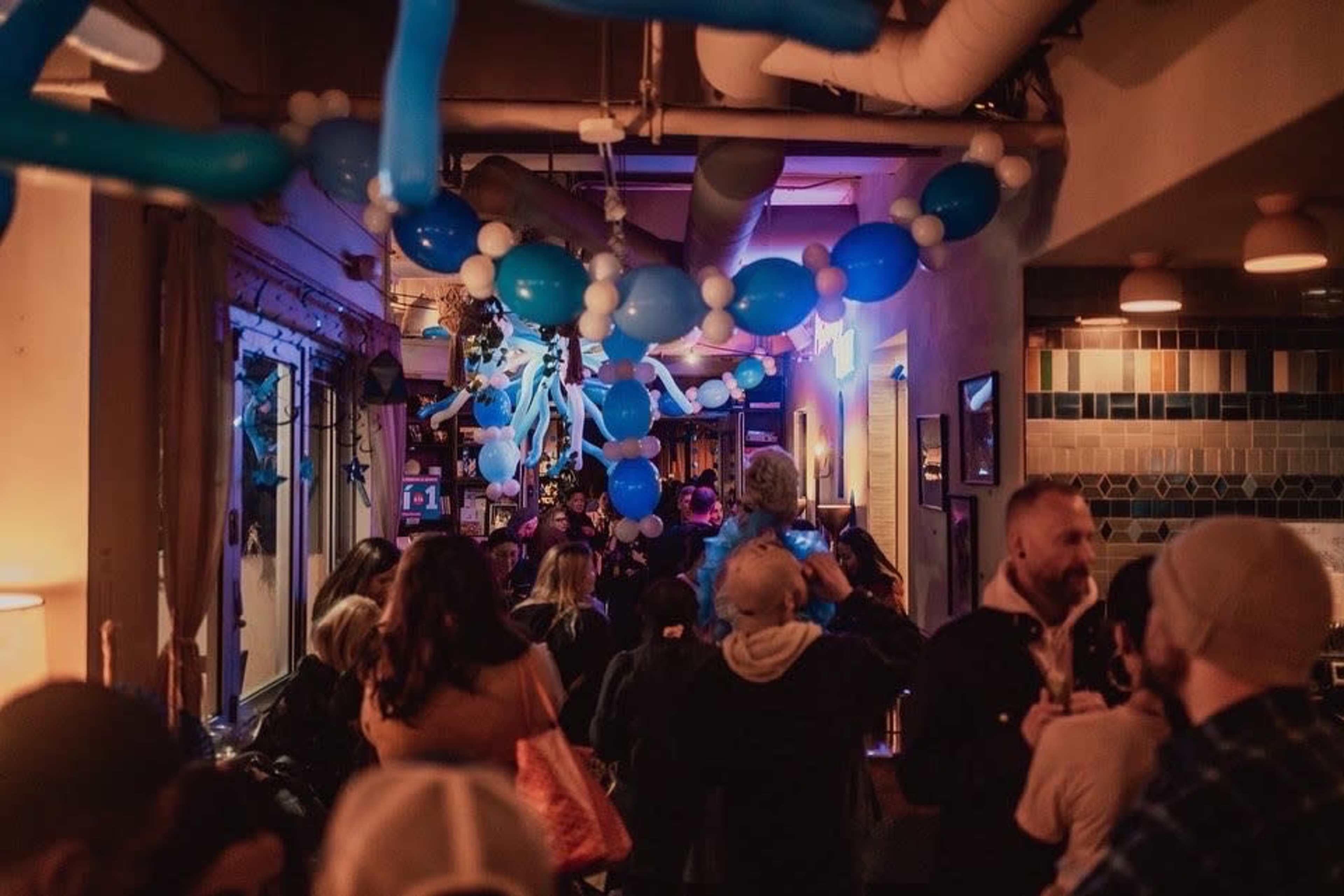 A crowded bar decorated with blue and white balloons during an evening event.