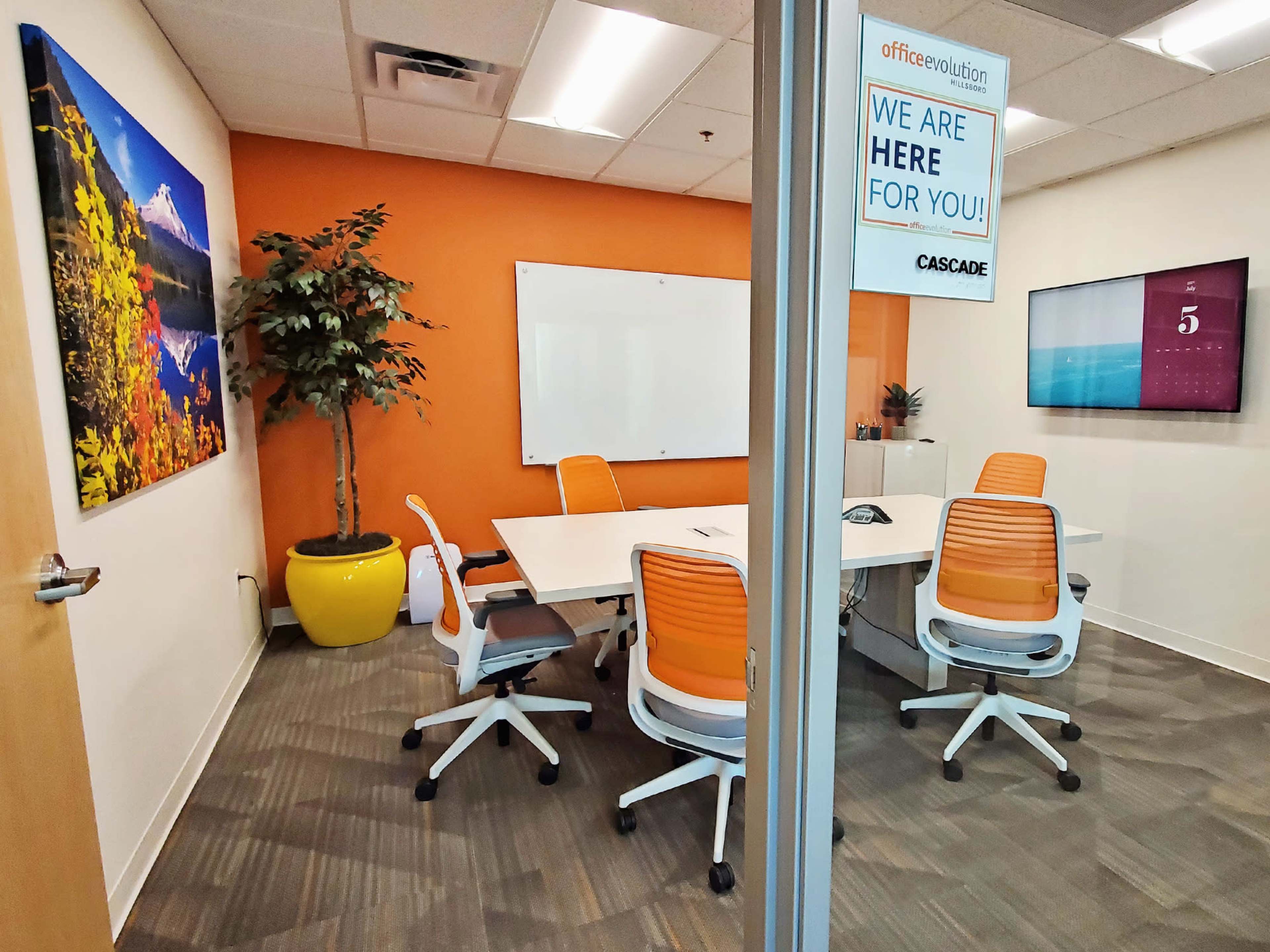 A modern conference room with a large table, orange-accented chairs, a whiteboard, a plant, and framed art on the walls.