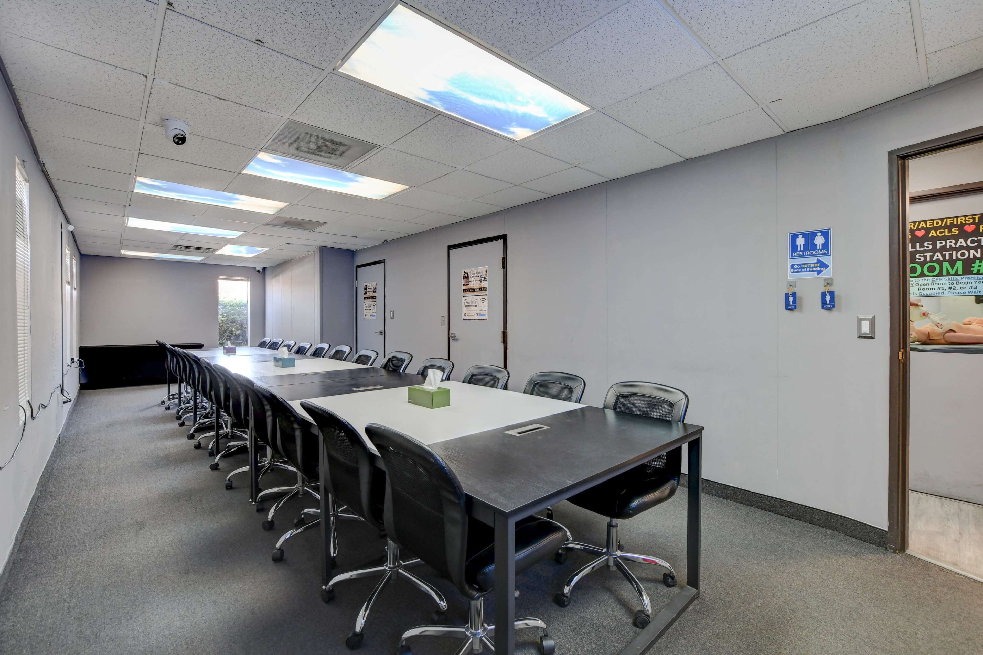 A long conference table with black chairs is positioned in a well-lit meeting room featuring a door labeled "Restroom" and large windows.