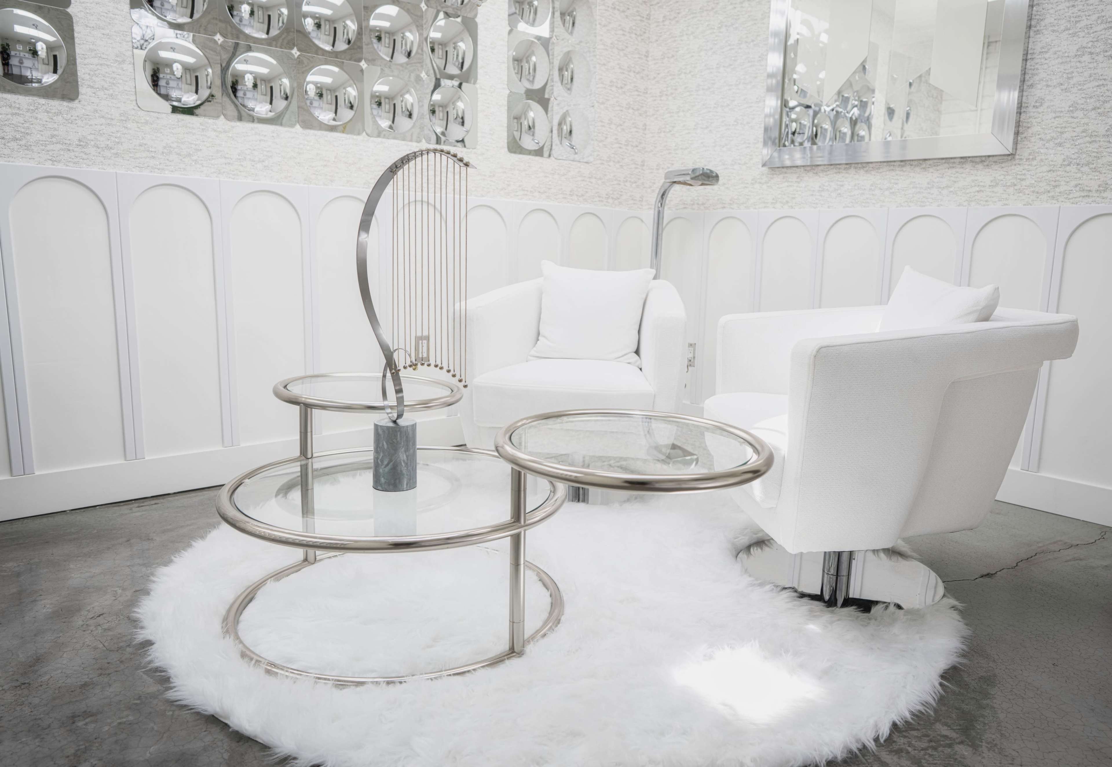 The image shows a modern seating area featuring white upholstered chairs, circular glass tables, and a fluffy rug against a light-colored wall with mirrored decorations.