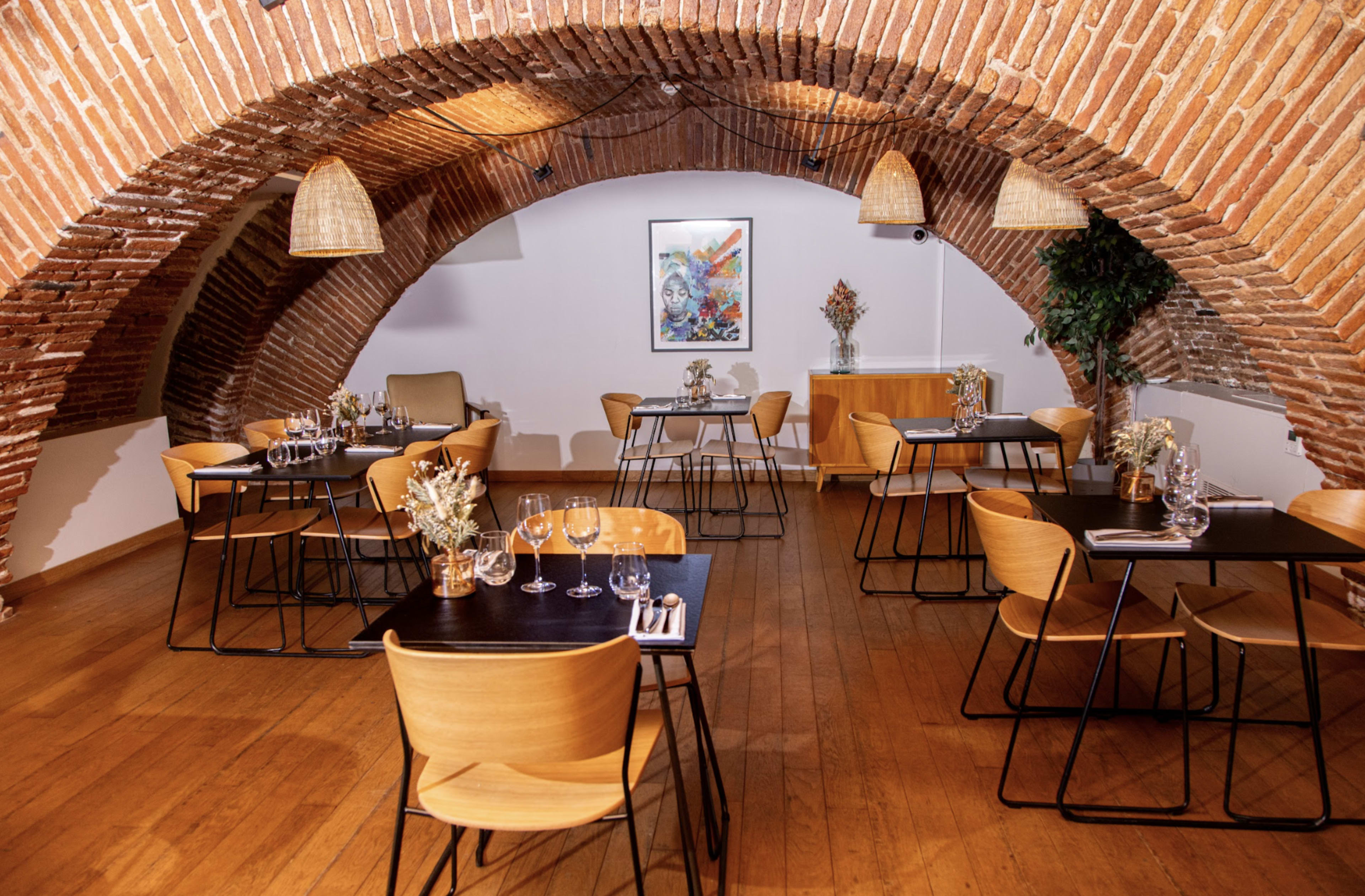 The image shows a restaurant interior with exposed brick arches, wooden floors, and tables set with glassware and floral centerpieces.