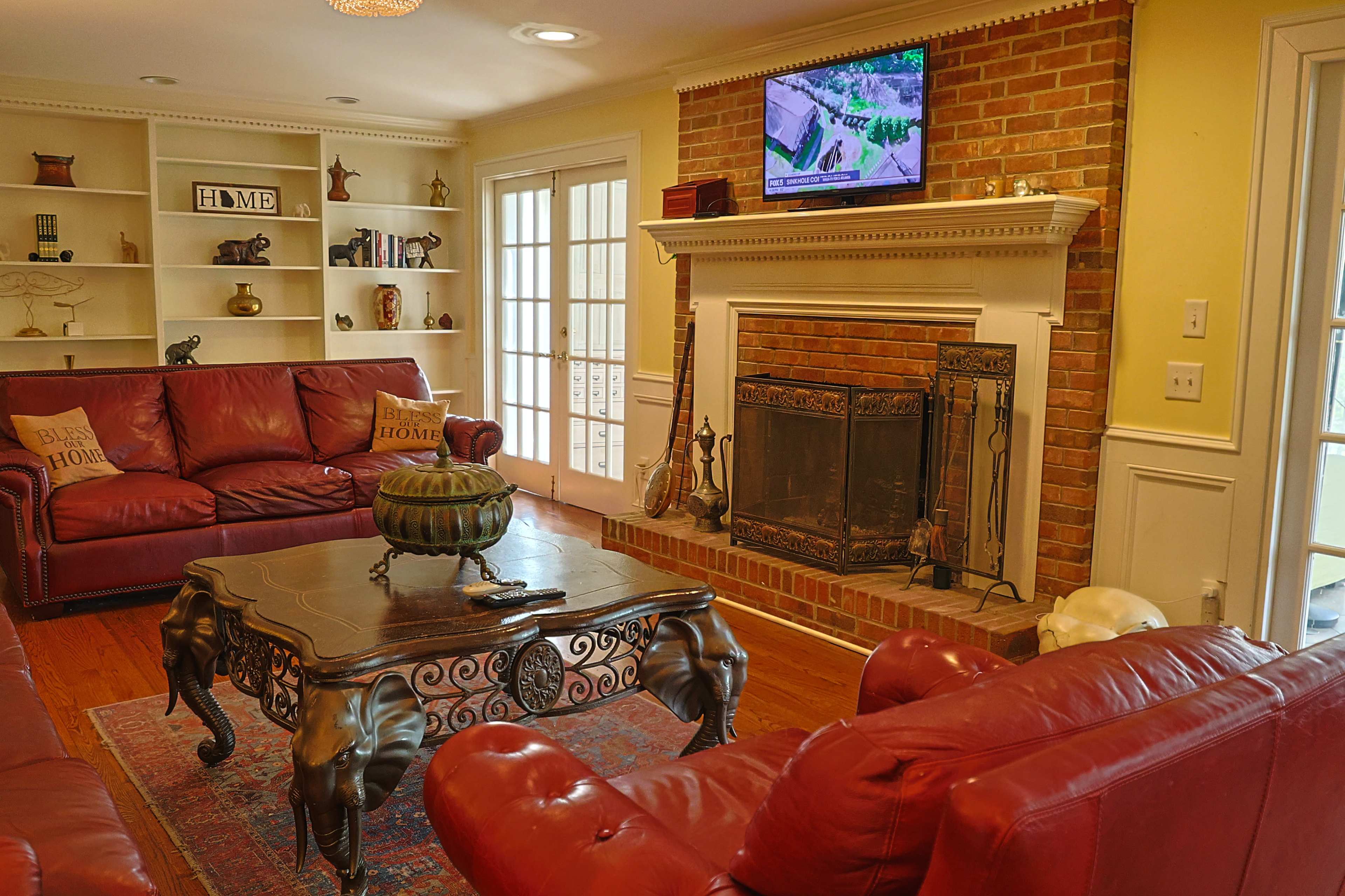 The living room features red leather sofas arranged around a wooden coffee table, with a brick fireplace and a television mounted above it.