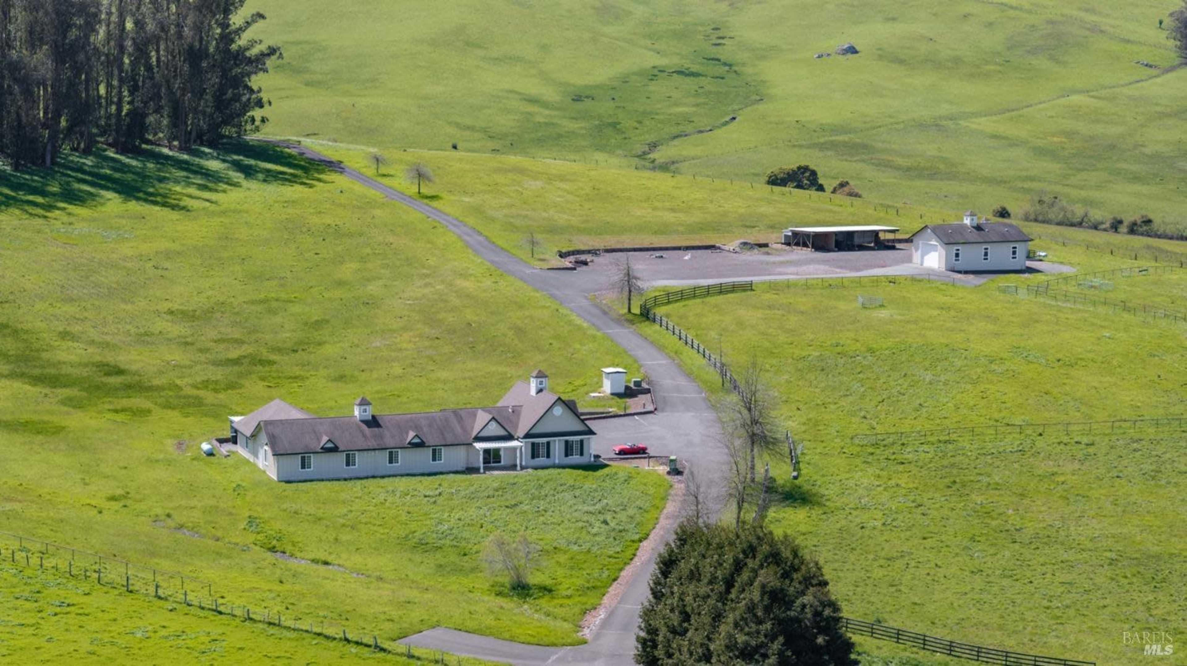 The image shows a spacious rural property featuring a large house with a dark roof, several outbuildings, and expansive green fields around it.