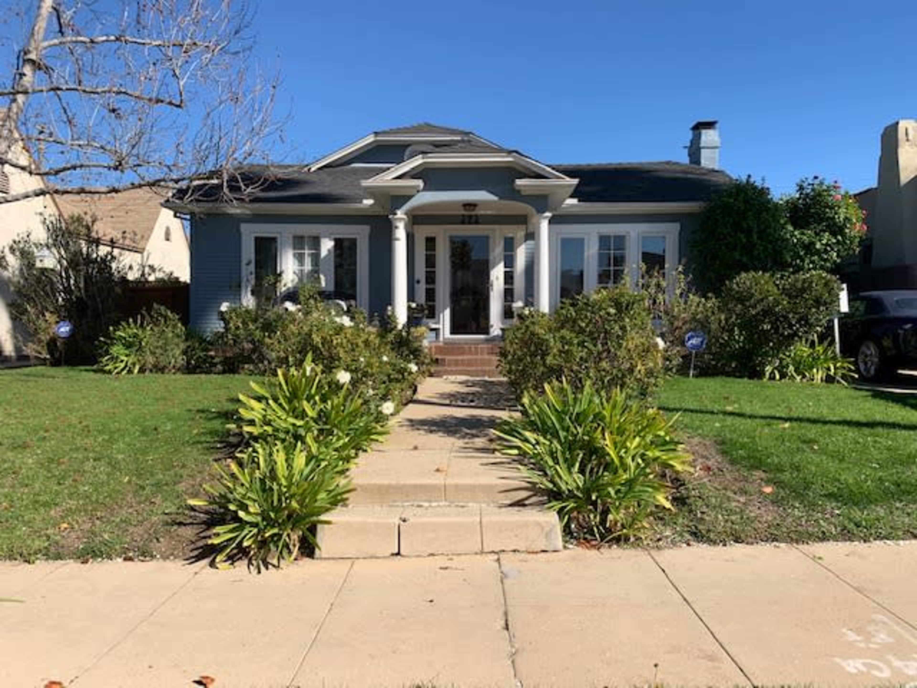 A single-story house with a front porch, surrounded by shrubs and a manicured lawn, is visible on a clear day.