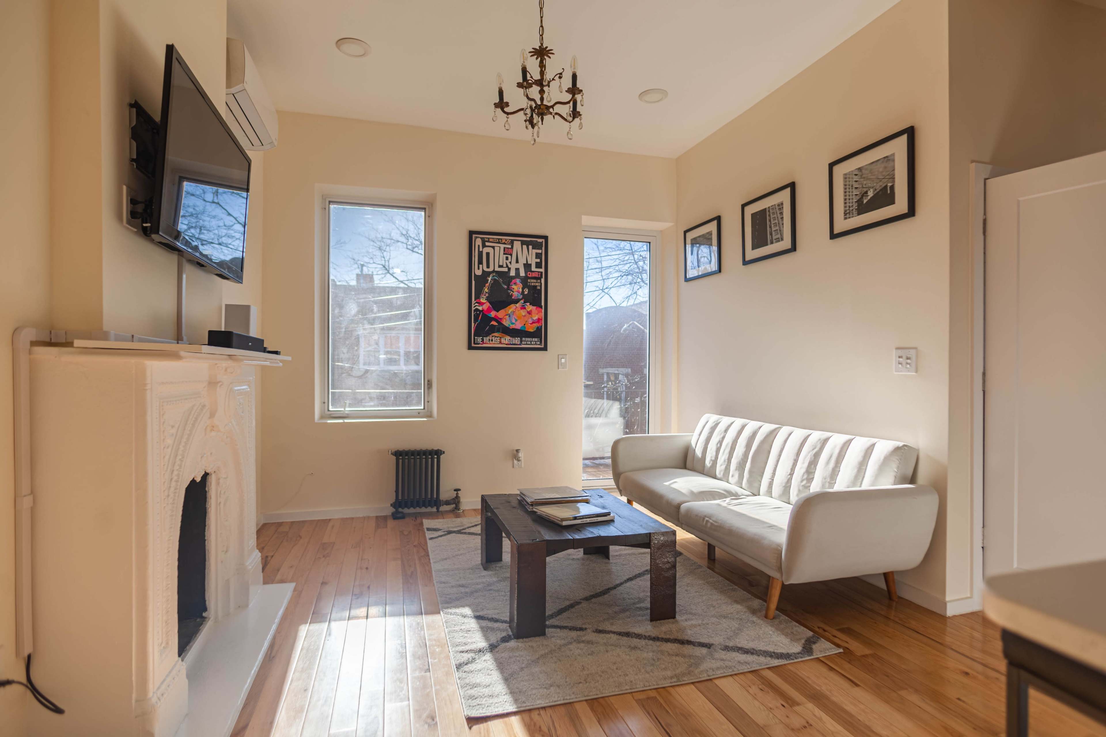 A cozy living room with a white sofa, a wooden coffee table, a framed poster on the wall, and large windows allowing natural light to enter.
