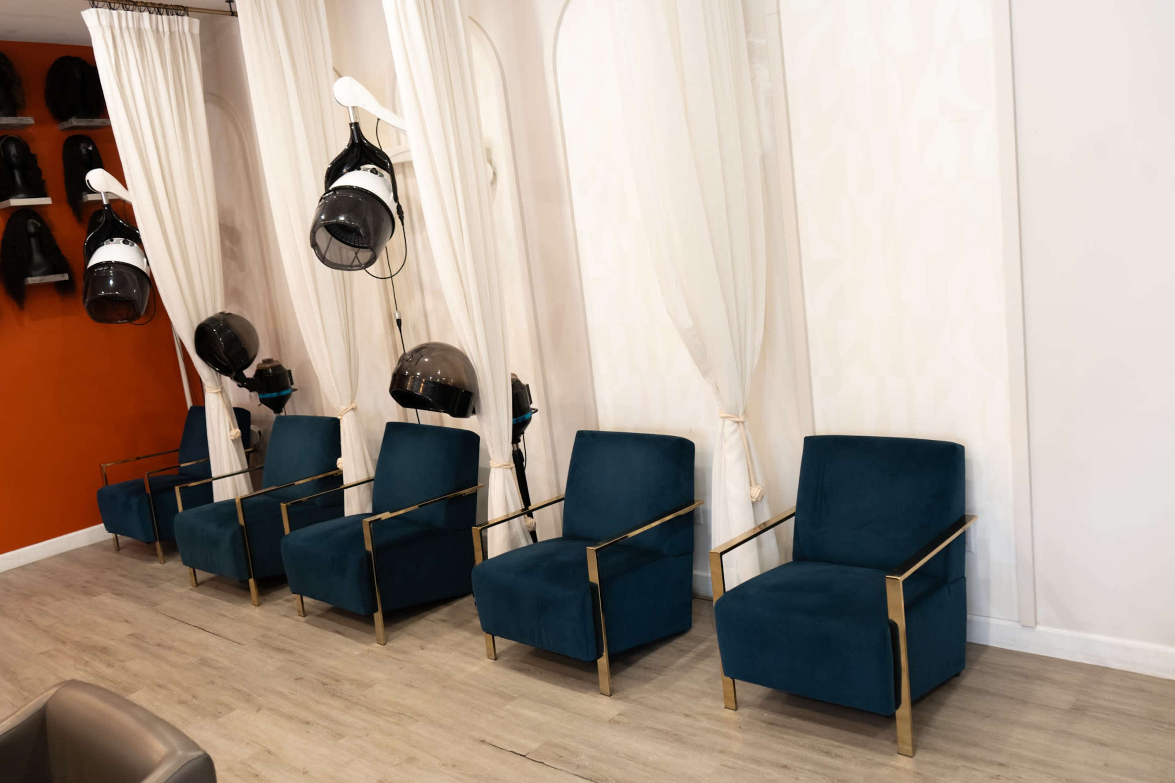 The image shows a hair salon interior with five blue armchairs arranged in a row, along with several hooded hair dryers hanging behind white curtains.