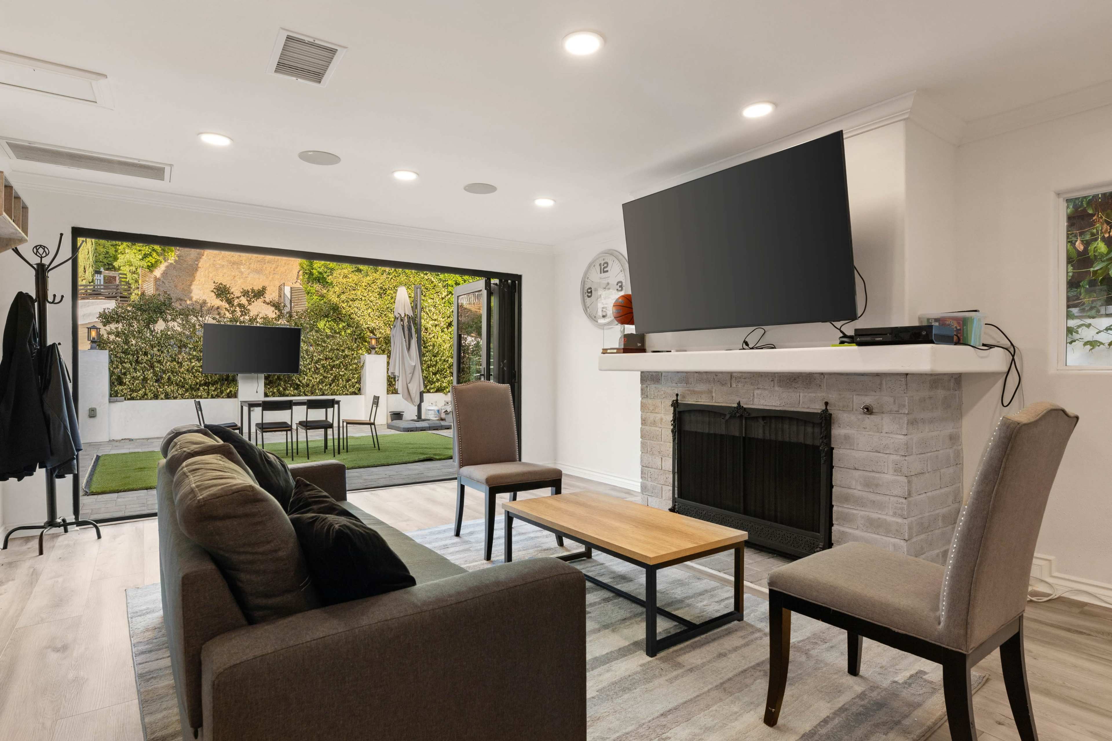 The image shows a modern living room with a dark sofa, two chairs, a coffee table, and a fireplace, leading to an outdoor patio through wide glass doors.