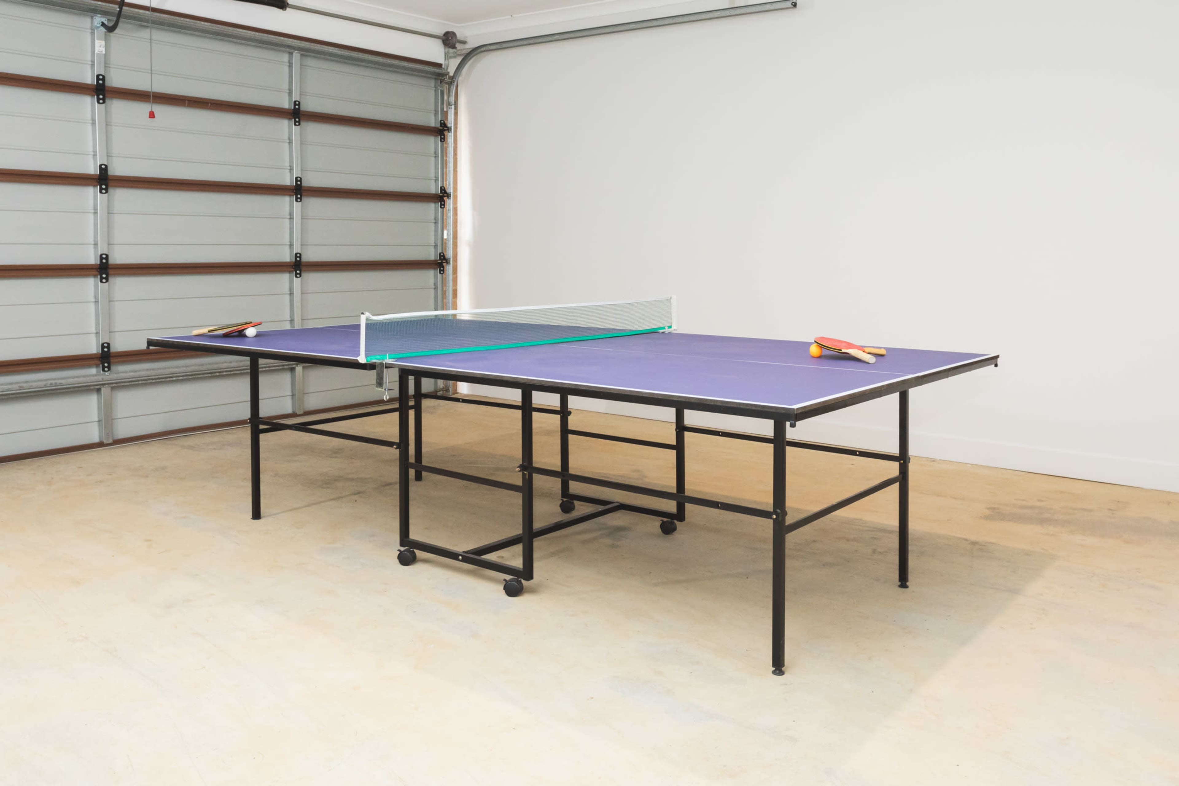 A ping pong table with paddles rests in a garage with a closed door.