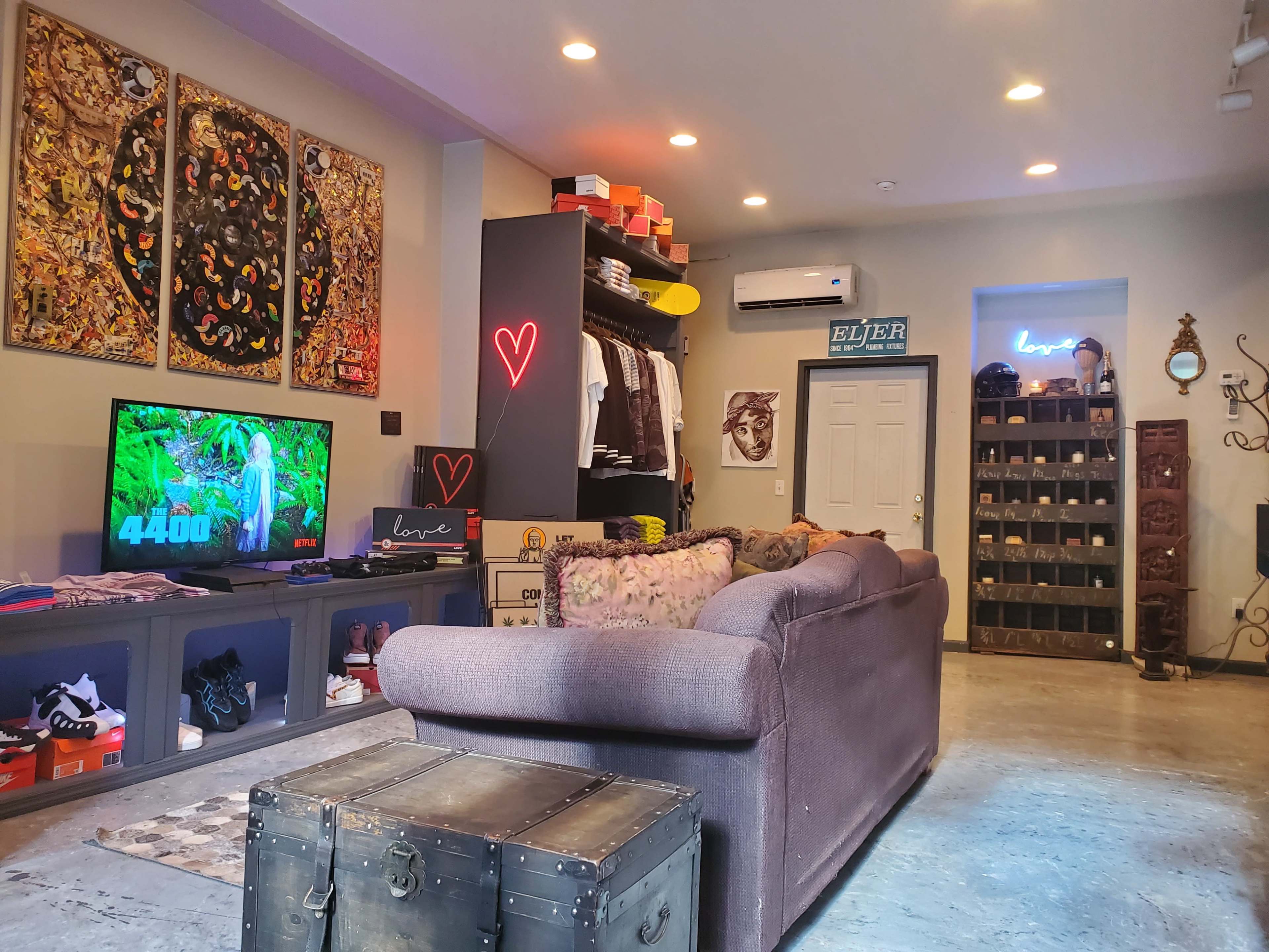 A stylish living room with a gray couch, a wall-mounted TV displaying a scoreboard, shelves filled with shoes and clothing, and decorative art pieces.