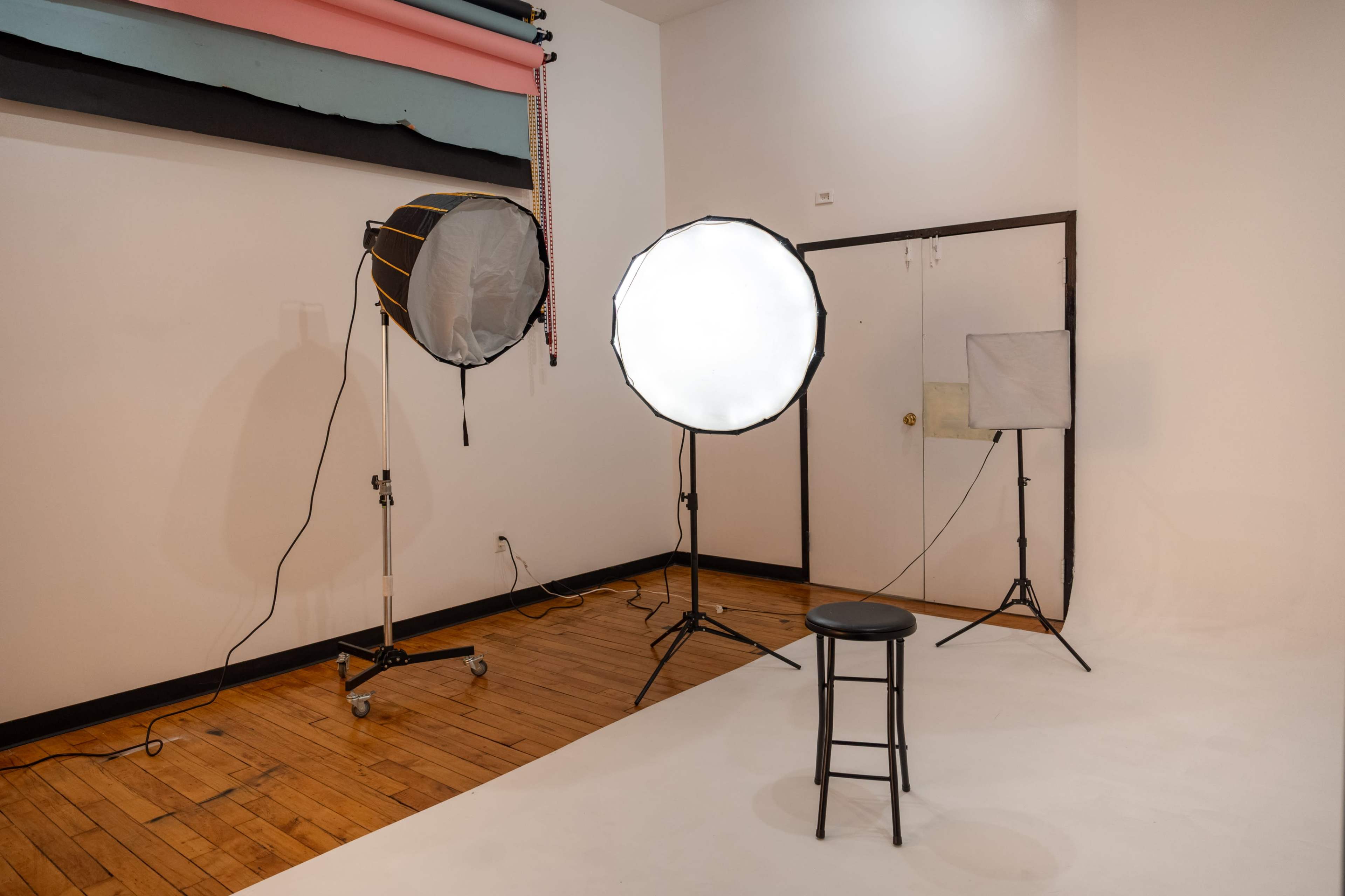 The image shows a photography studio with a stool in the center, two softbox lights, and a backdrop setup.