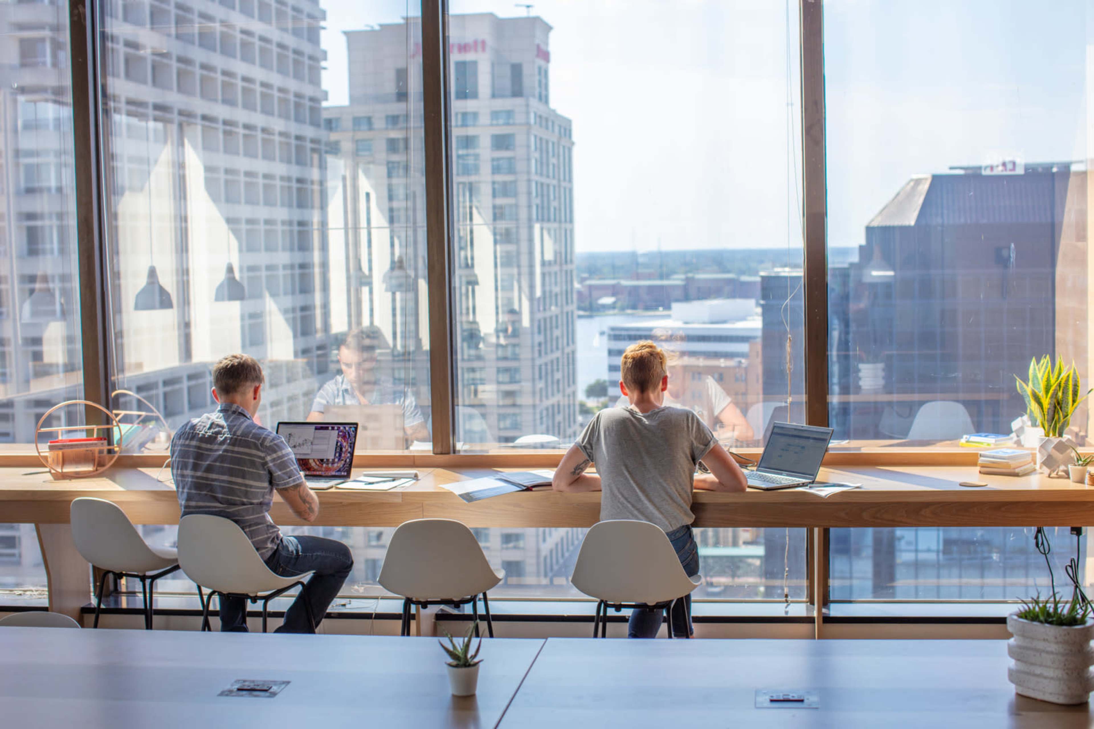 Two people work on laptops at a wooden table next to large windows overlooking a cityscape.