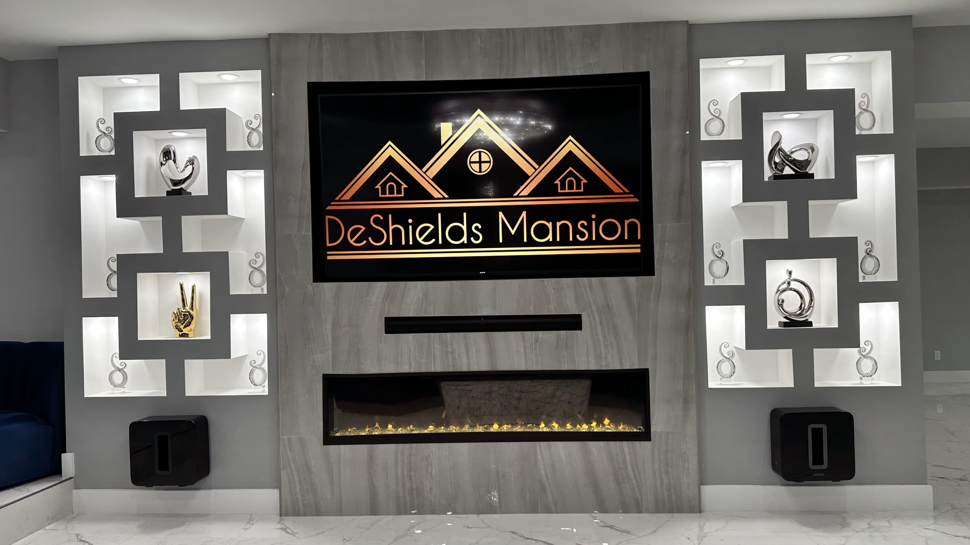 A modern living room wall featuring a large TV with the "DeShields Mansion" logo centered above a decorative fireplace, flanked by illuminated shelves displaying artistic pieces.