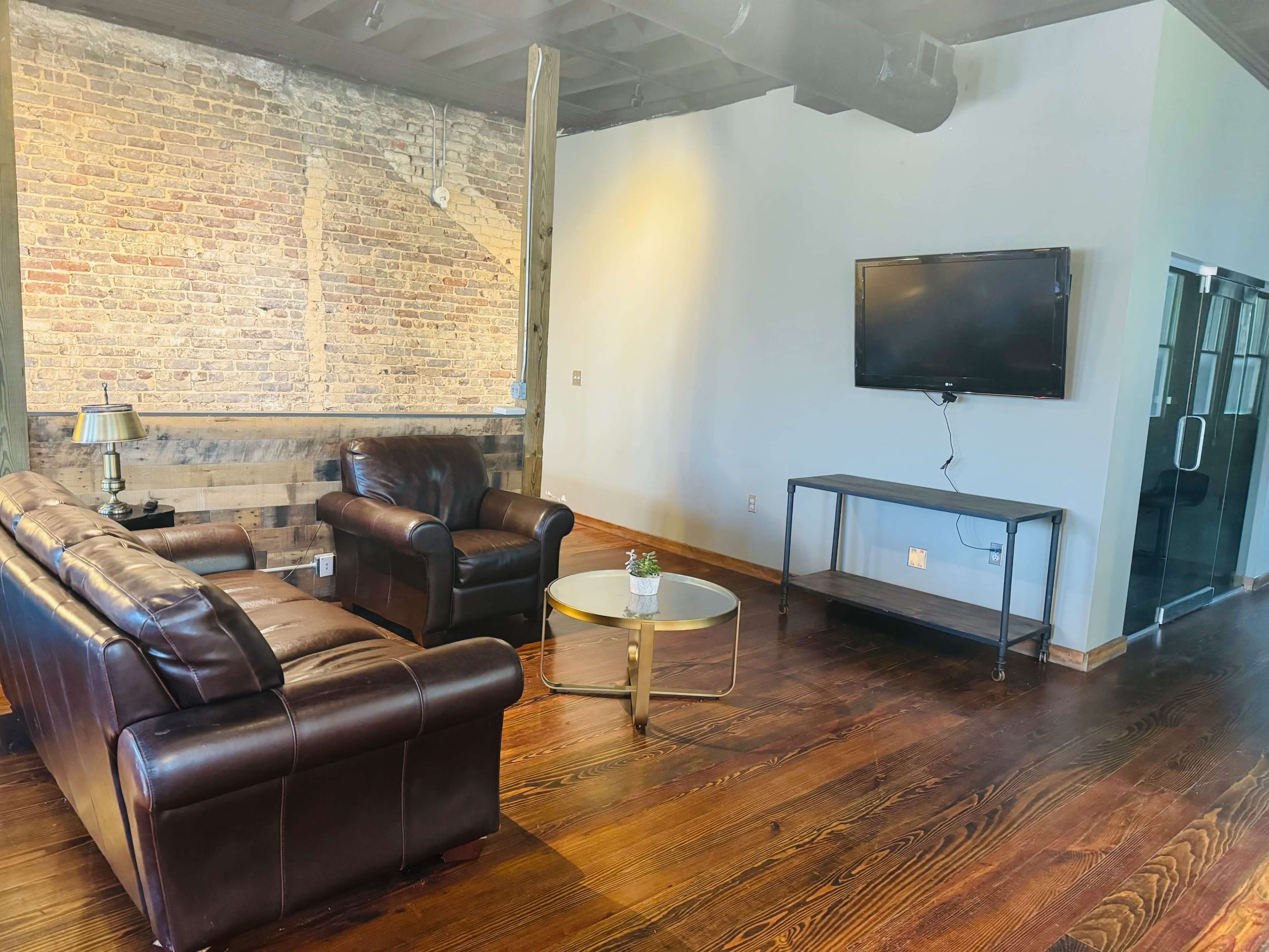 A modern living room with a leather sofa set, a glass coffee table, and a wall-mounted television, against a backdrop of exposed brick and wooden beams.