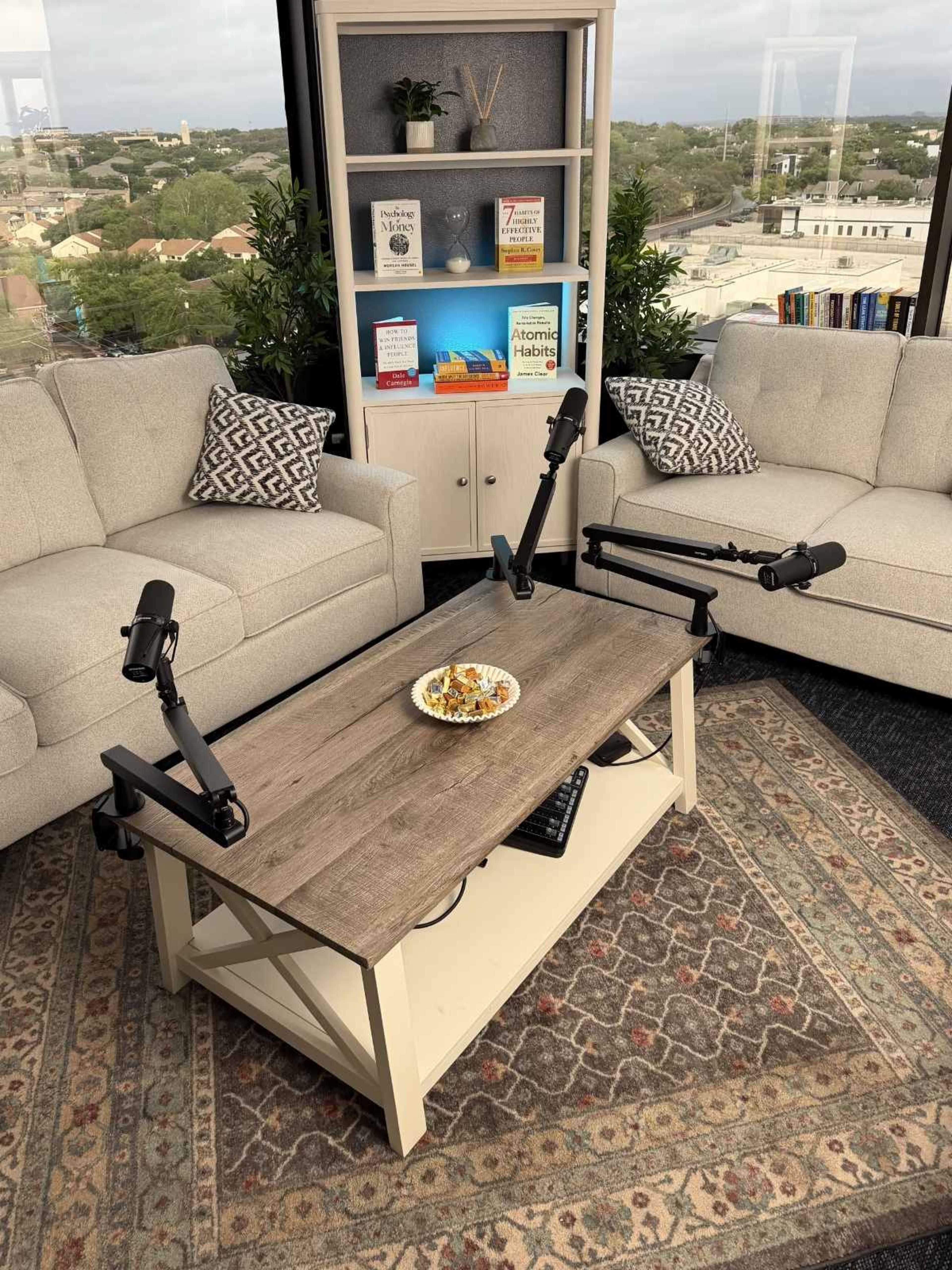 The image shows a modern sitting area with two light-colored sofas facing a wooden coffee table that has two microphone stands and a plate of food, all set against a backdrop of a bookshelf filled with books.