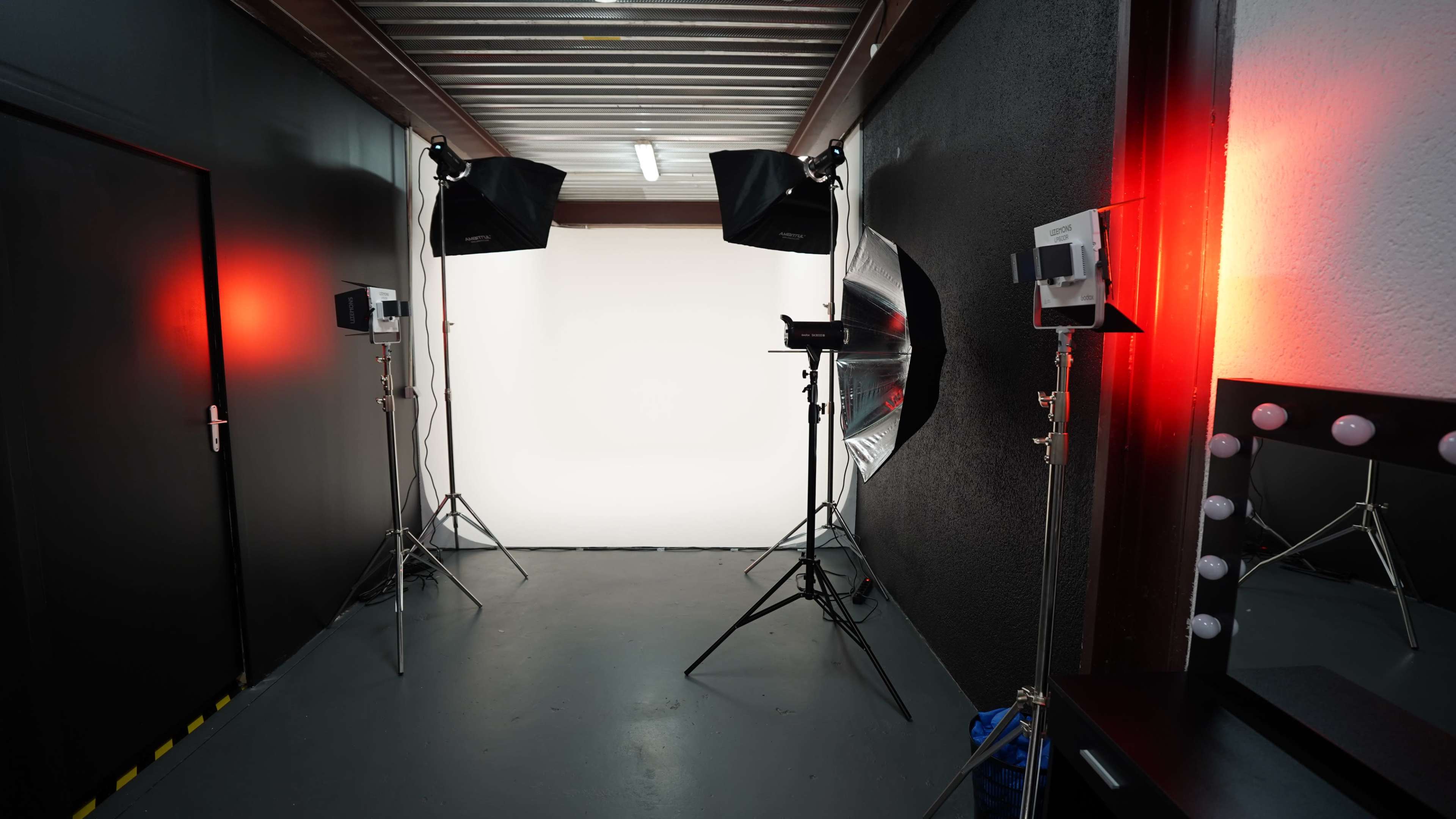 The image shows a photography studio with black walls, lighting equipment on stands, and a white backdrop.