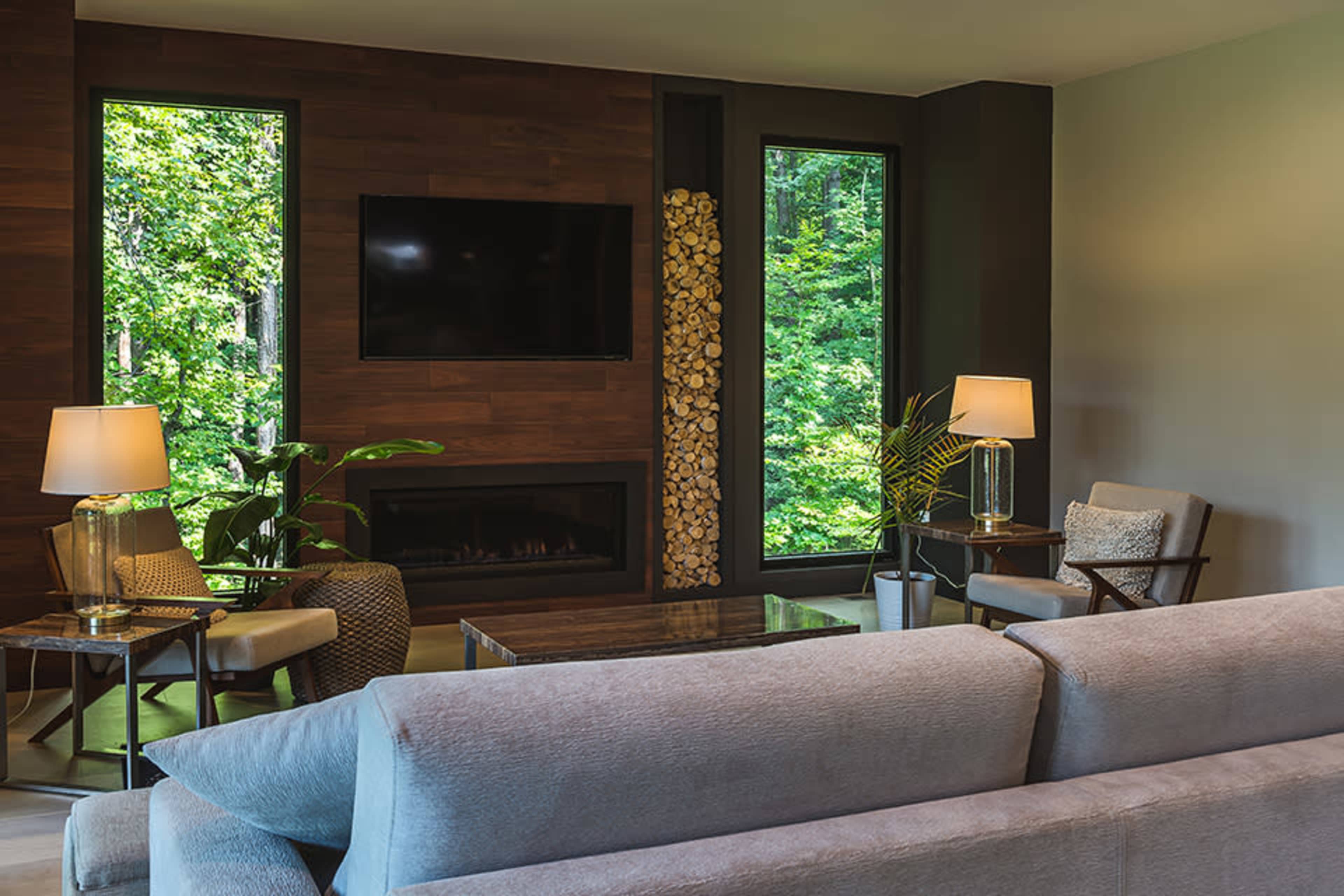 A cozy living room features a modern gray sofa, two chairs, a large flat-screen TV mounted on a wooden wall, and large windows offering a view of lush greenery.