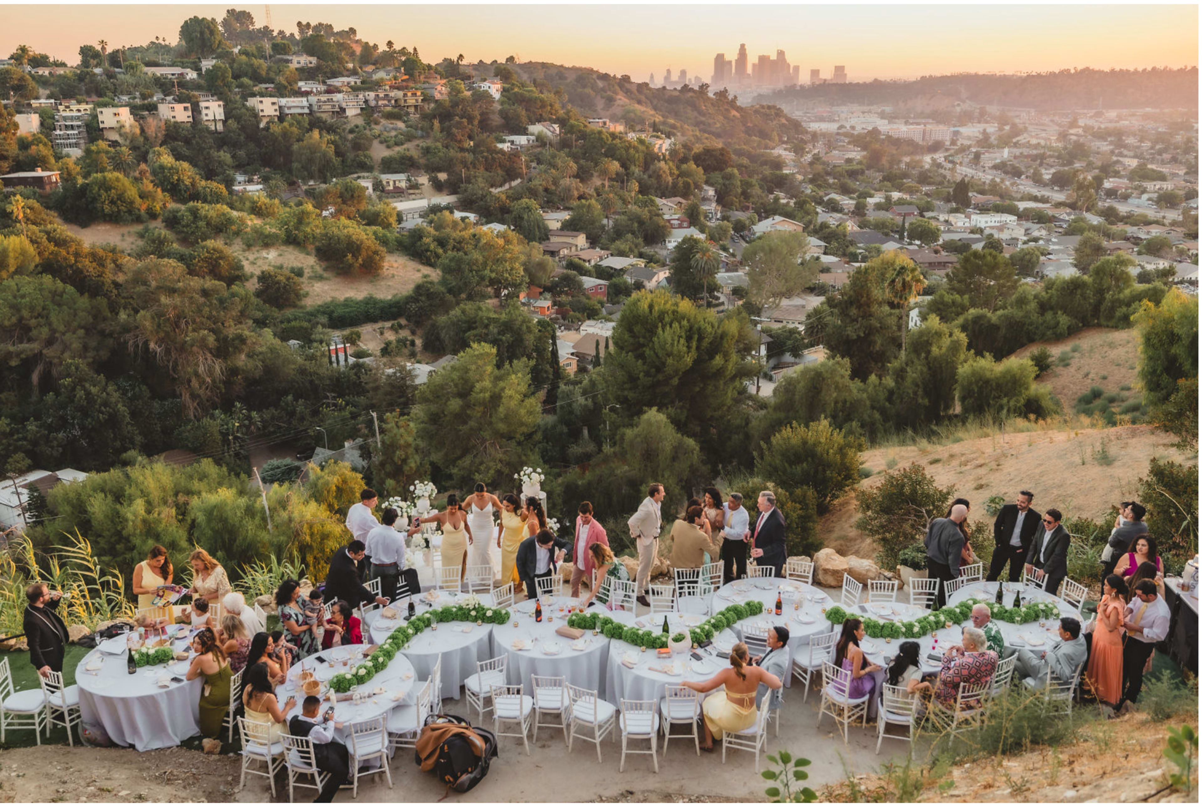 A large outdoor wedding reception is set up on a hillside with round tables arranged for guests, overlooking a view of the city skyline at sunset.
