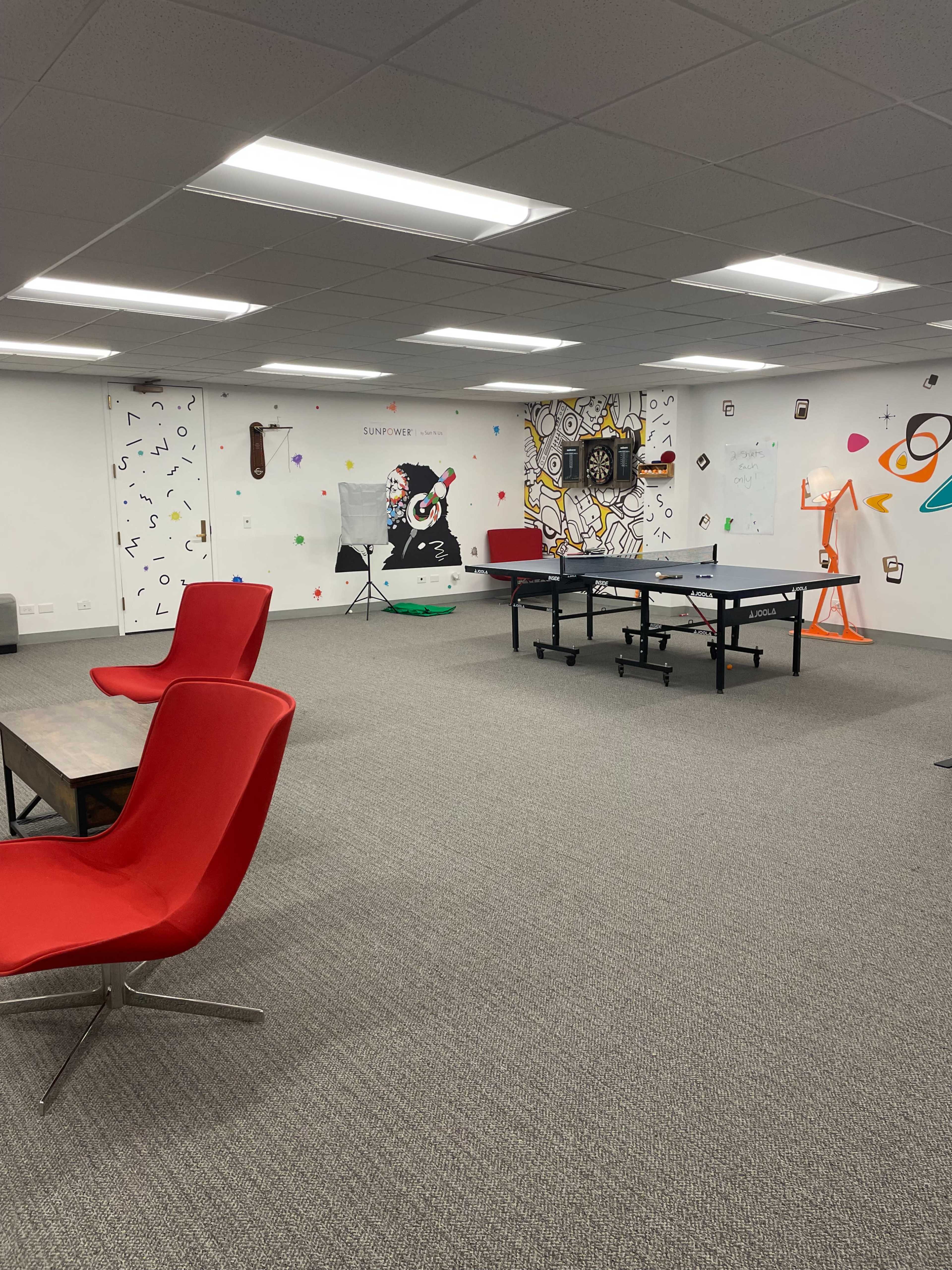 The image shows a spacious office or recreational area featuring a ping pong table, red seating, and colorful murals on the walls.