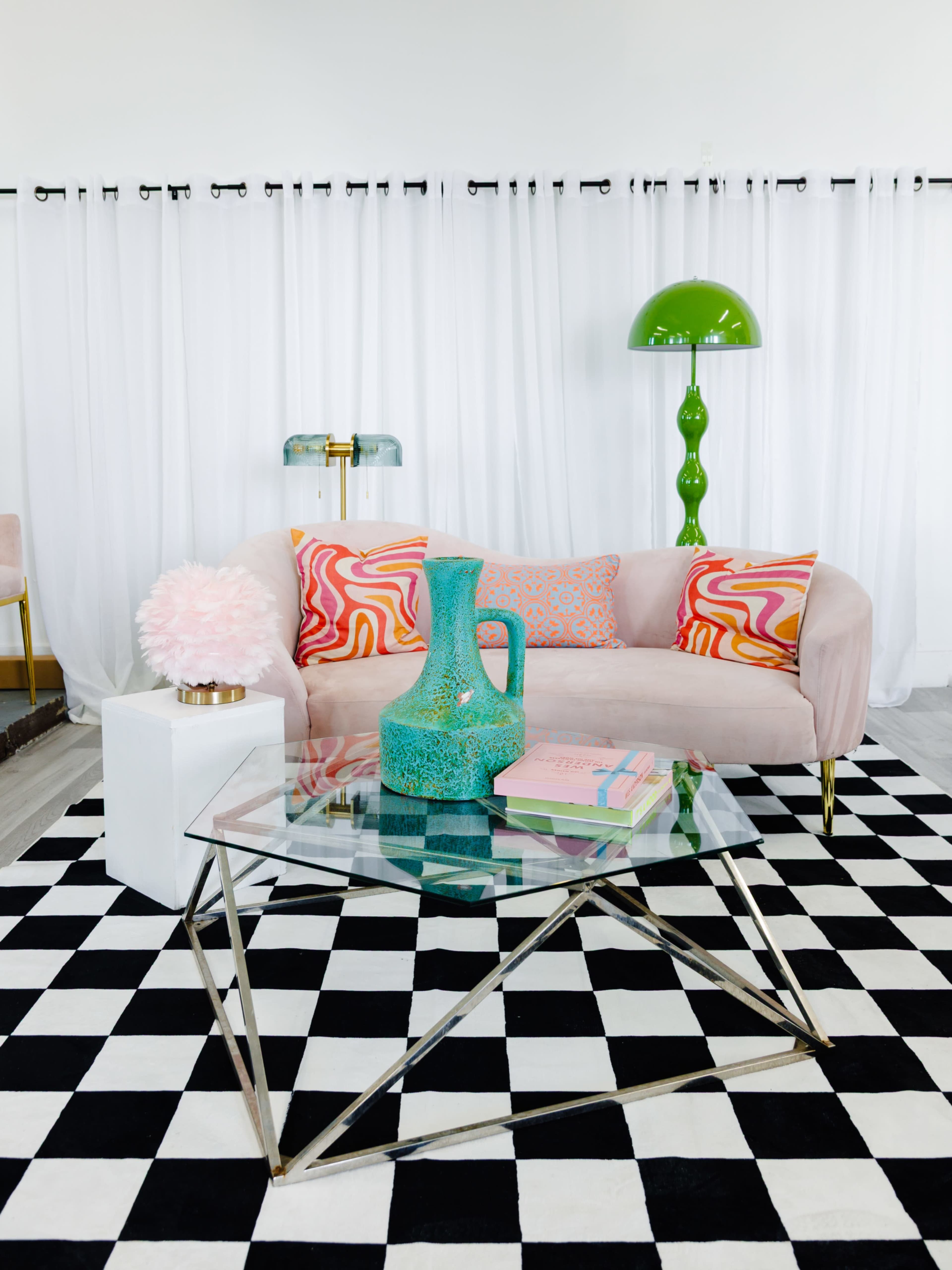 A pink couch with patterned cushions sits on a black and white checkered rug, alongside a glass coffee table and a green lamp, with white curtains in the background.