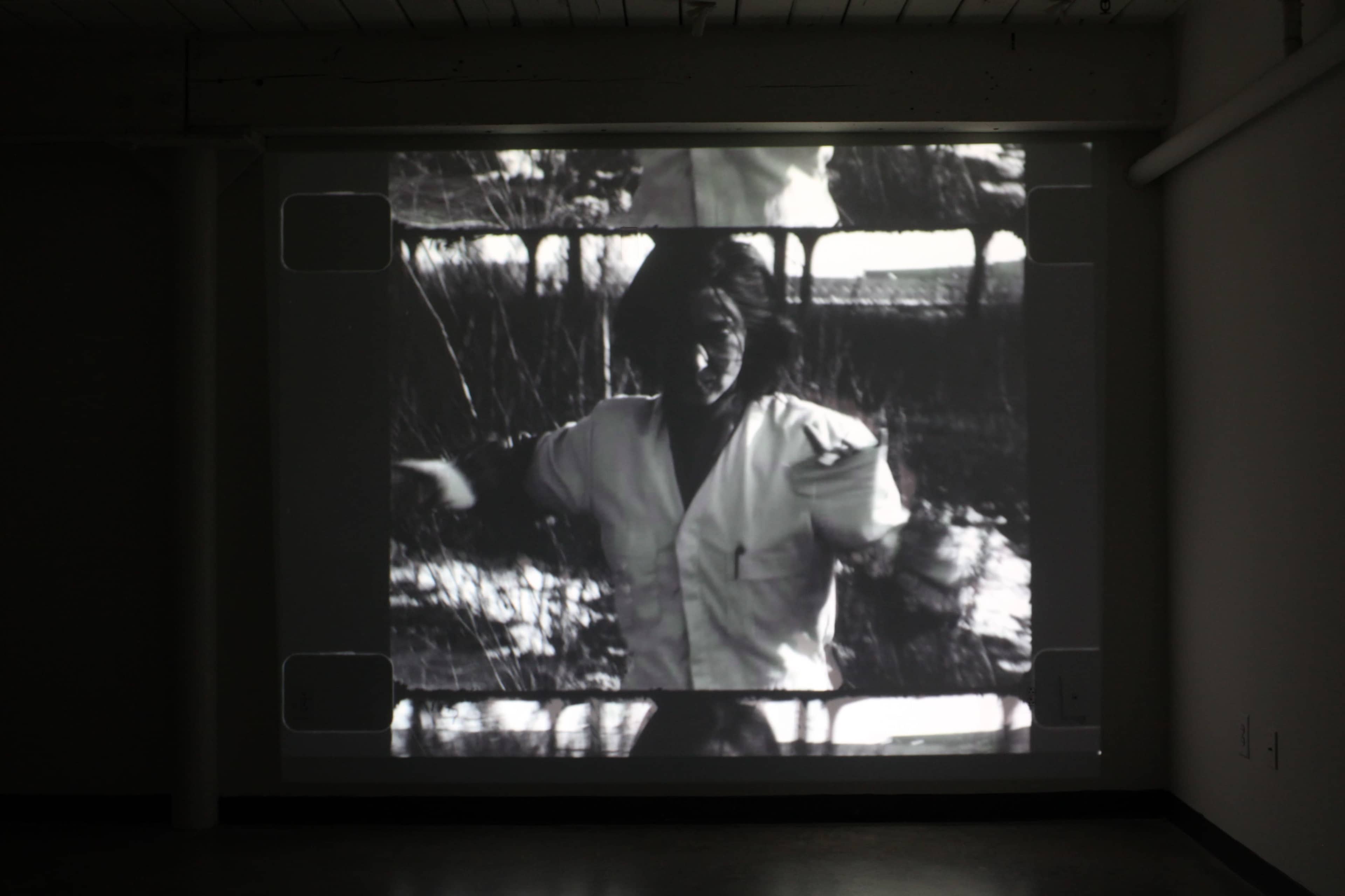 The image shows a black and white film projection of a figure moving in a natural setting, displayed on a wall in a dimly lit room.