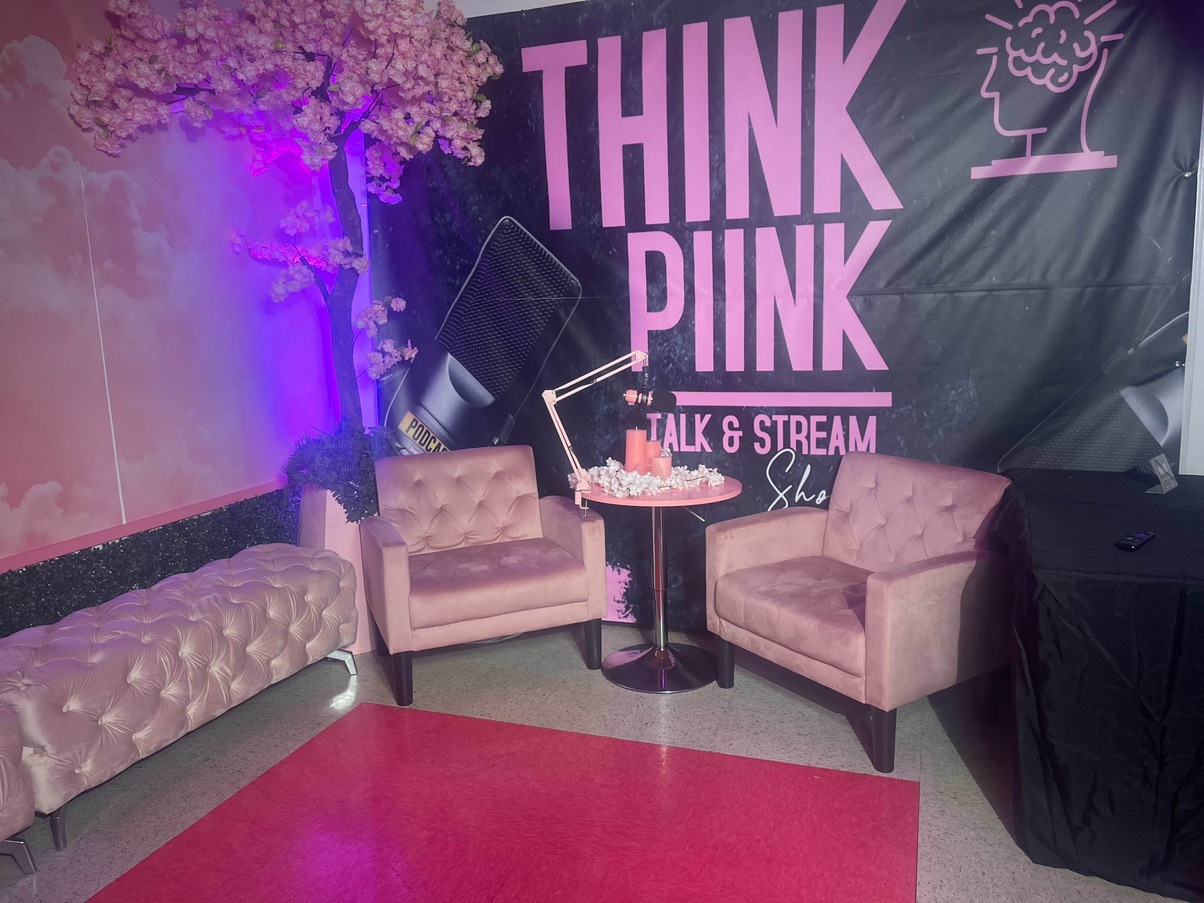The image shows a decorated area featuring two pink armchairs positioned around a small table, with a backdrop displaying "THINK PINK TALK & STREAM" and artificial cherry blossom trees.