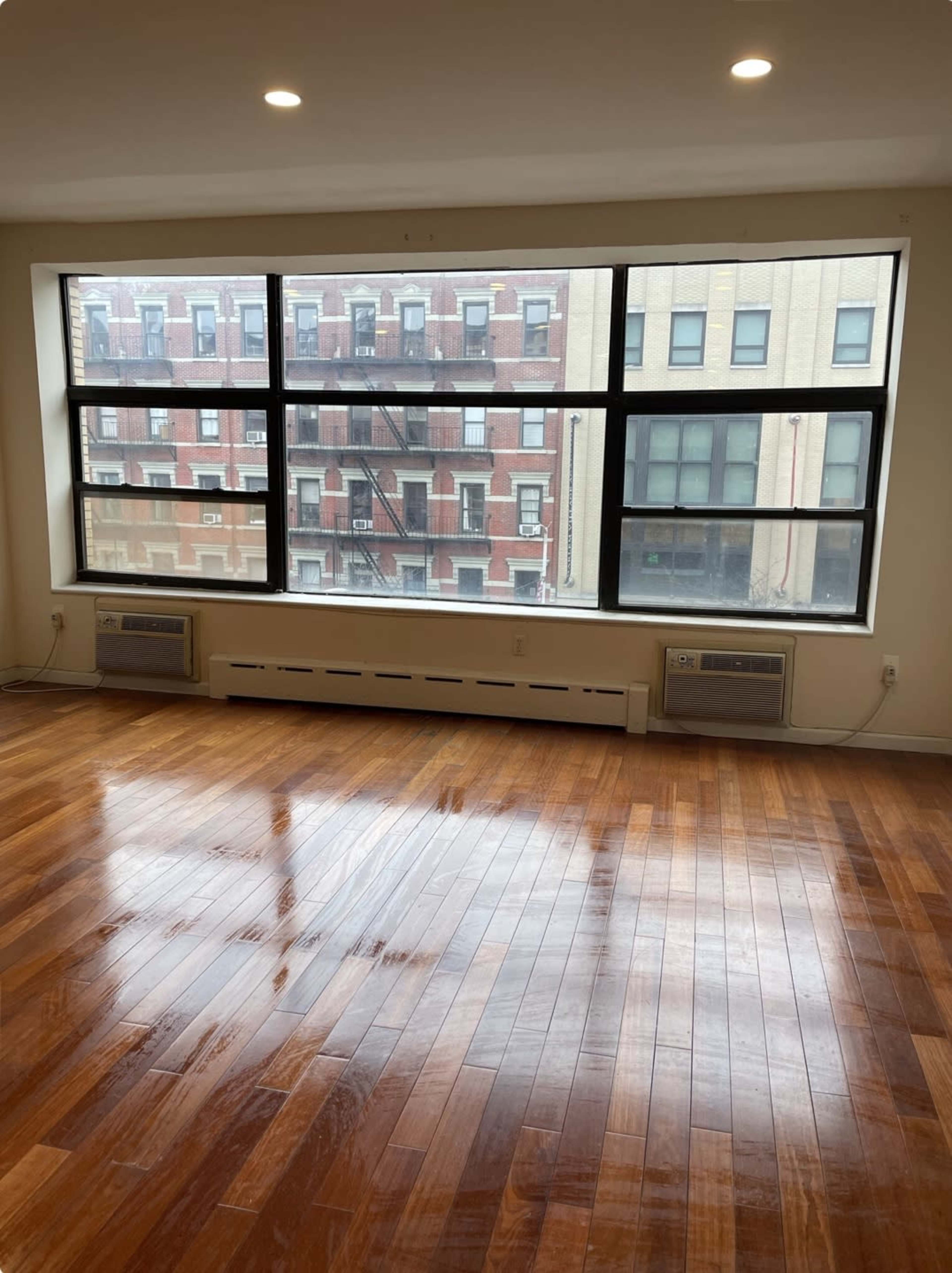 A room with polished wooden floors and large windows overlooking a neighboring brick building.