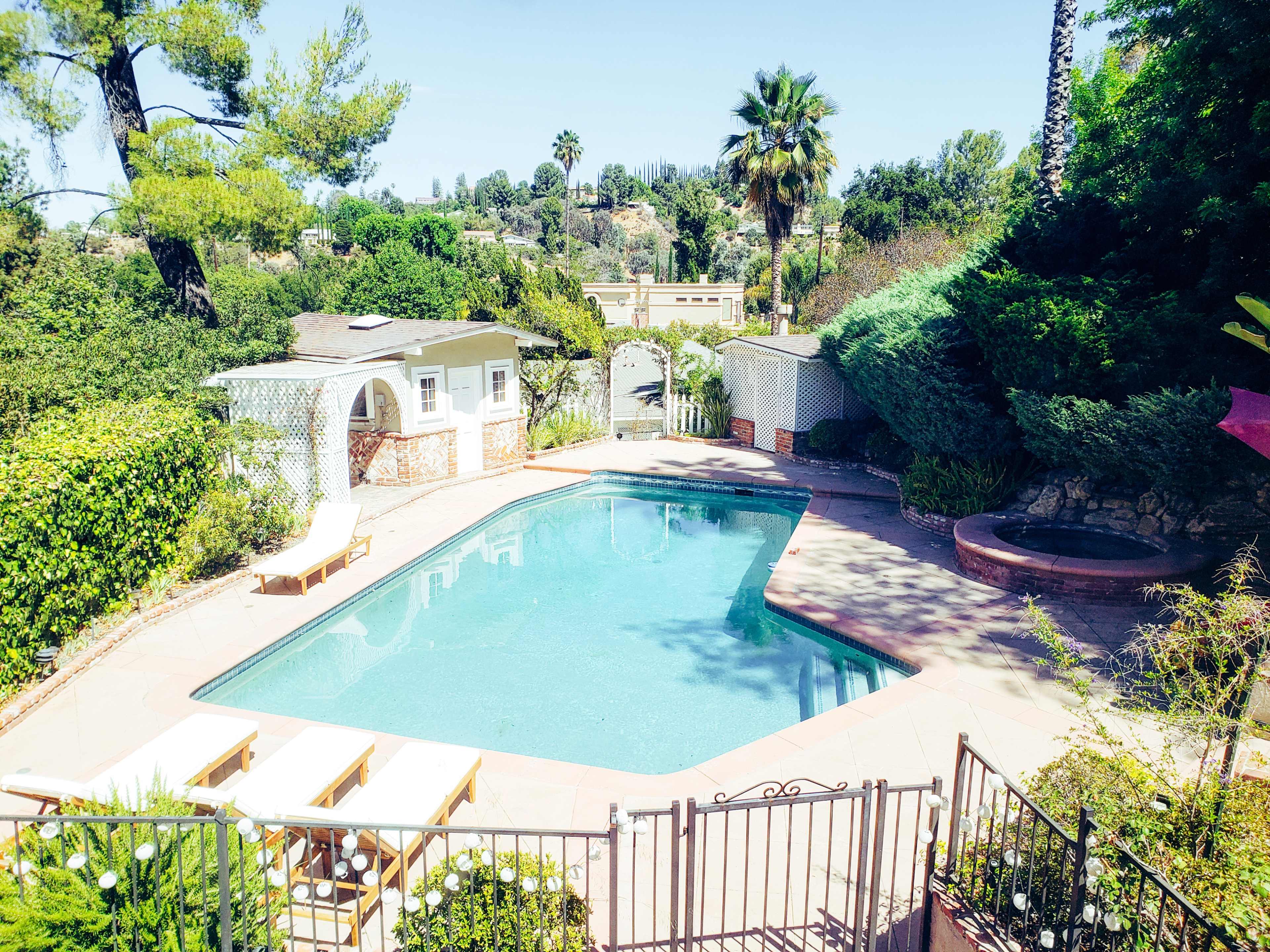 The image shows a swimming pool surrounded by greenery and outdoor lounge chairs, with a small gazebo in the background.