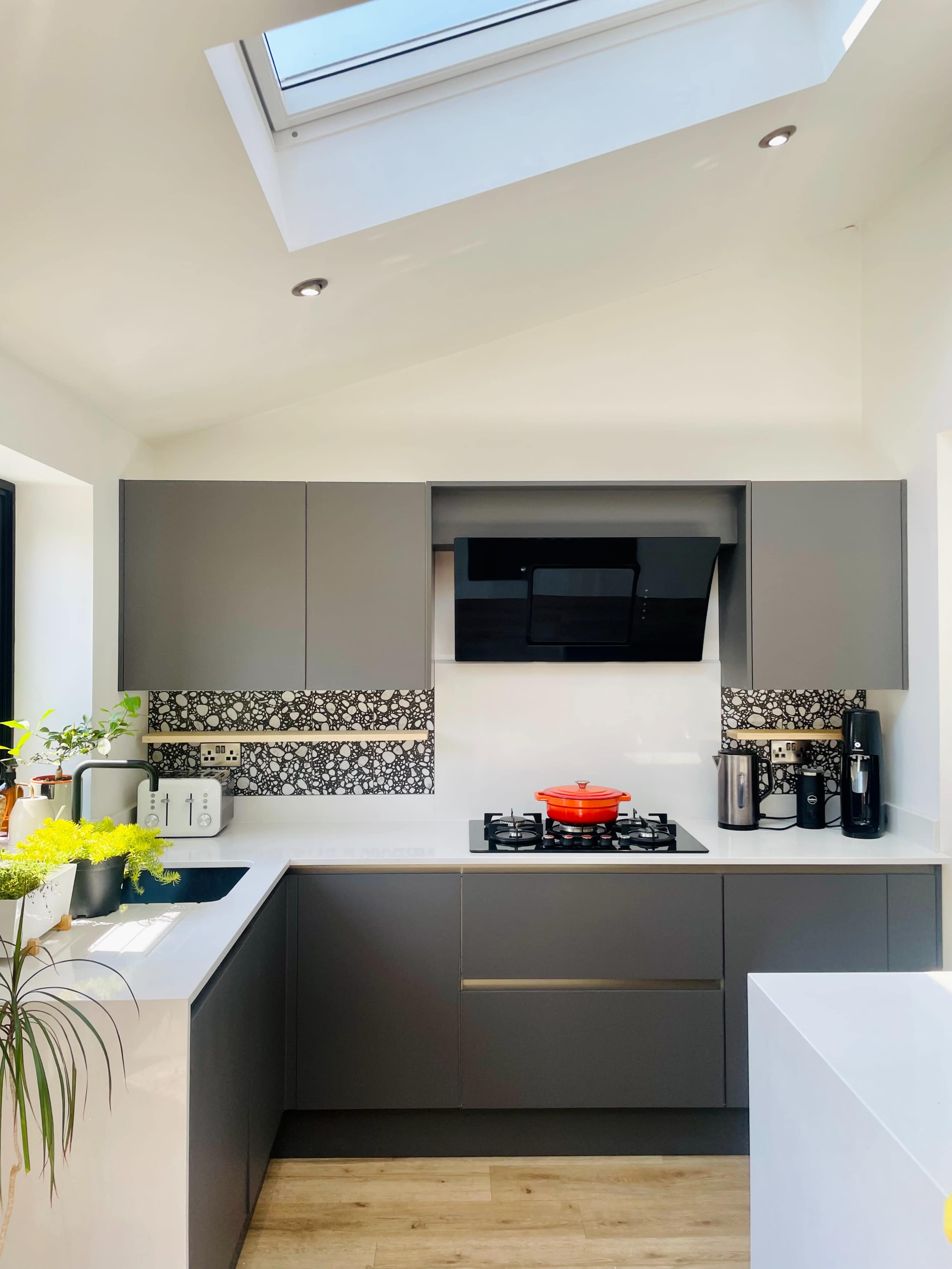 A modern kitchen features gray cabinetry, a black wall-mounted hood, and a red pot on the stove, with a skylight allowing natural light.