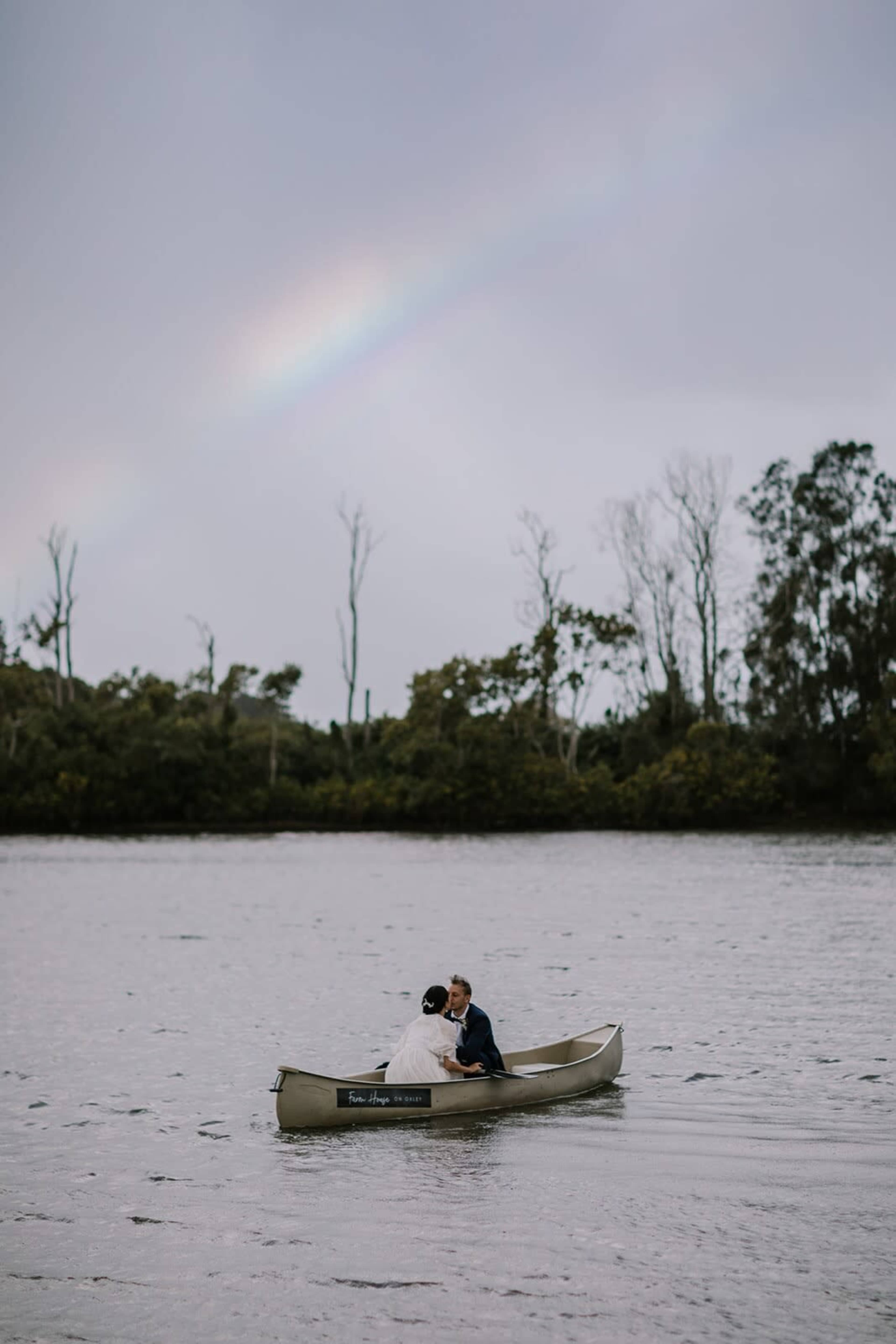 A couple in wedding attire is kissing in a canoe on a calm body of water under a rainbow.
