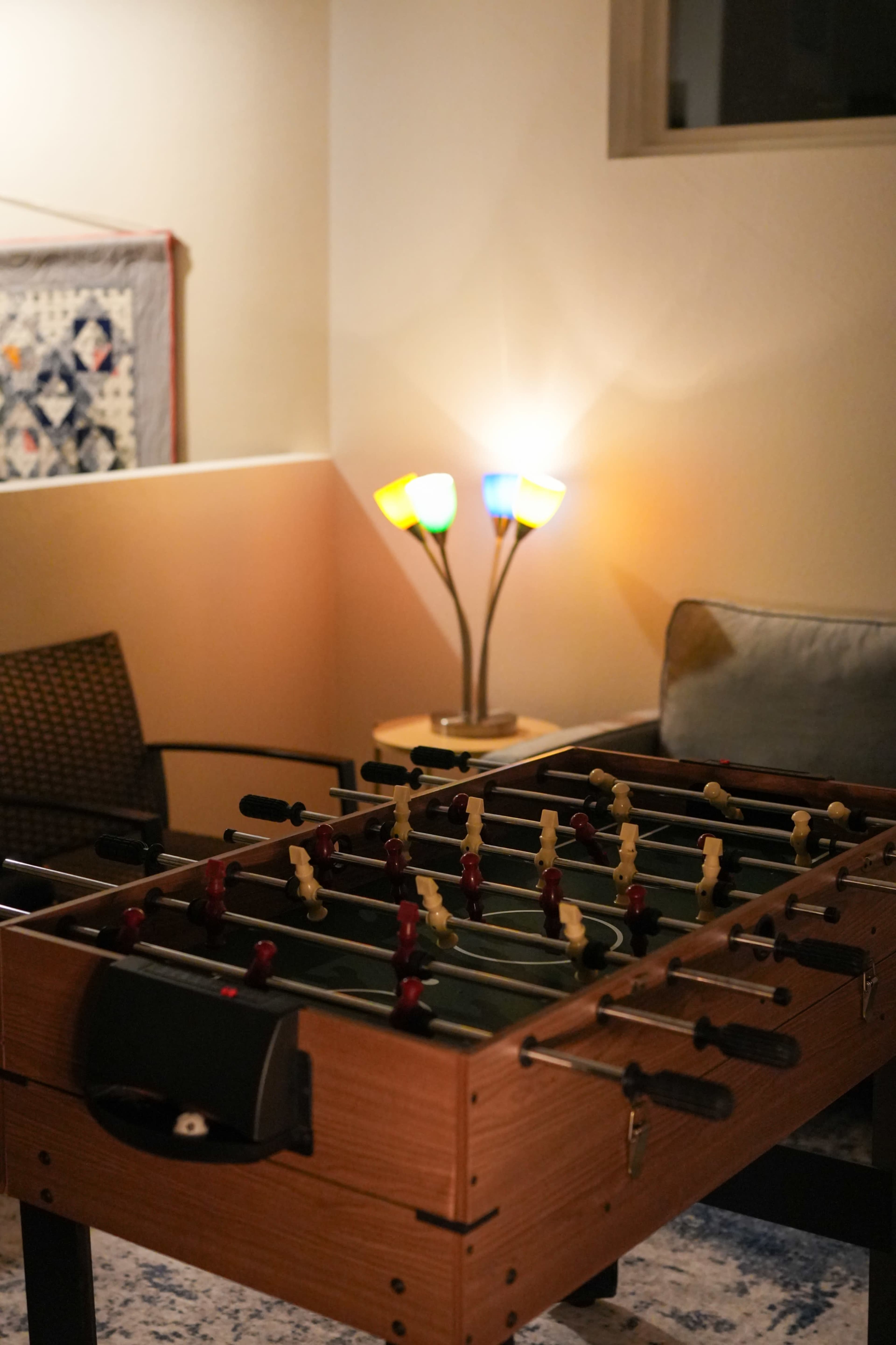 A foosball table is positioned in a cozy room illuminated by a colorful lamp, with a seating area in the background.