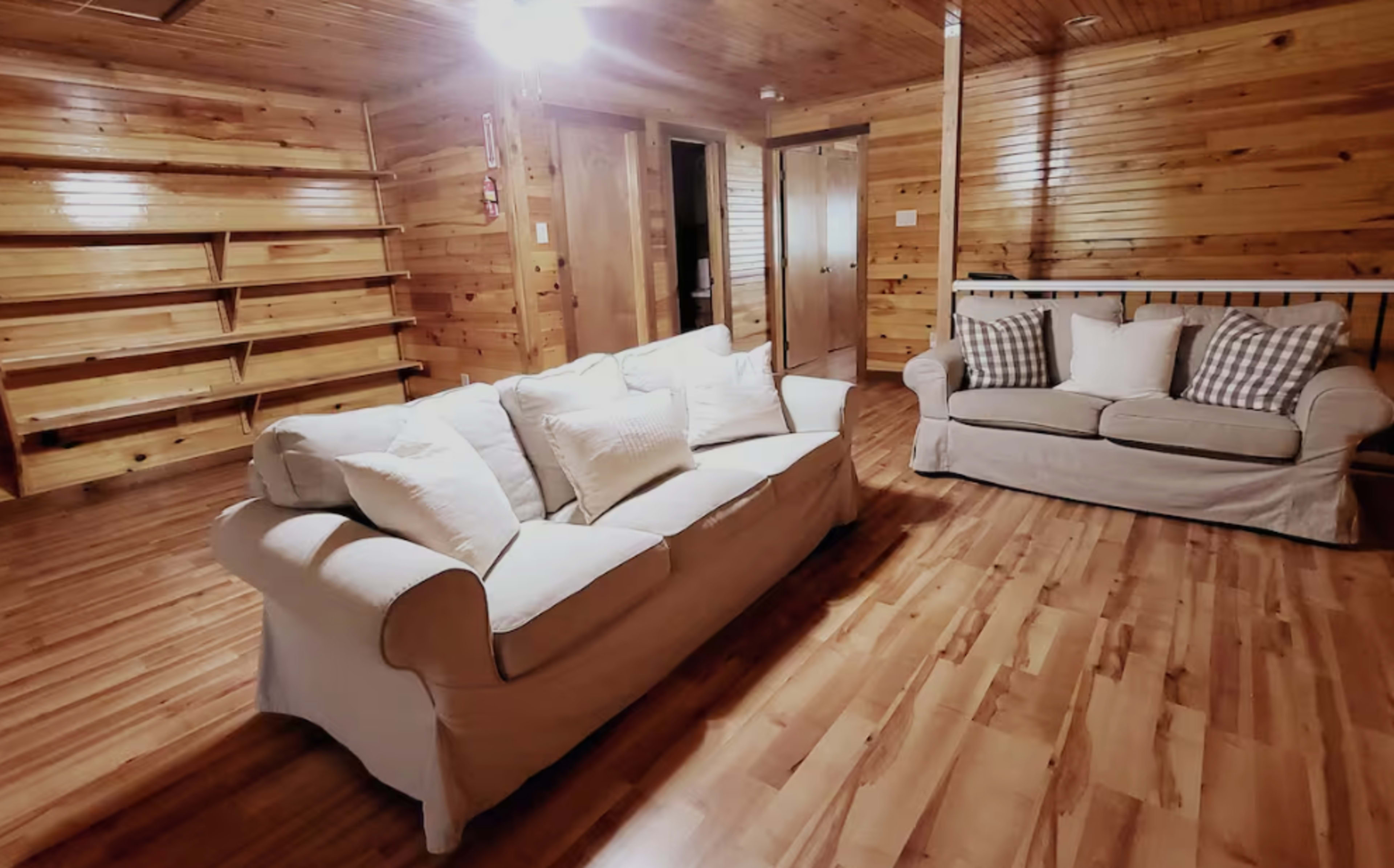 The image shows a cozy interior room with wooden walls and floors, featuring a light-colored couch with cushions and a set of shelves along one wall.
