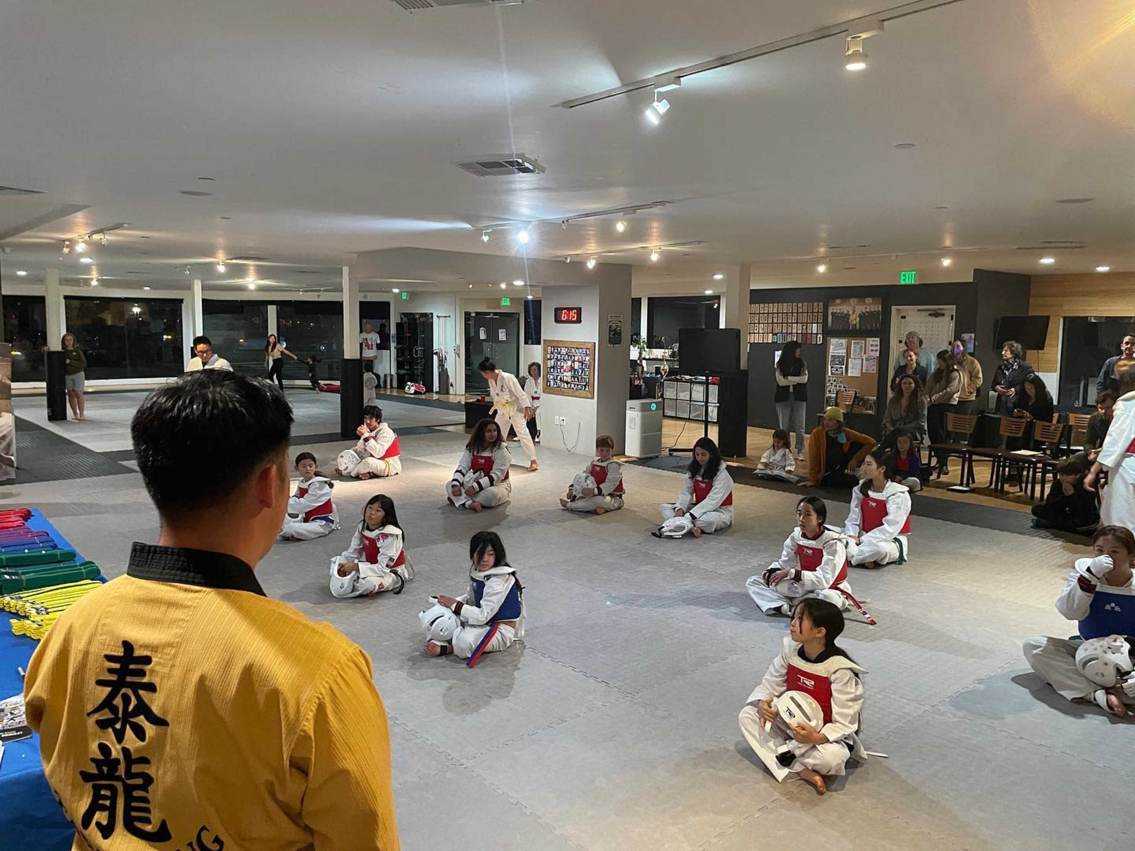 A karate instructor stands in front of a group of children in white uniforms, sitting in a dojo setting with mats and training equipment around them.