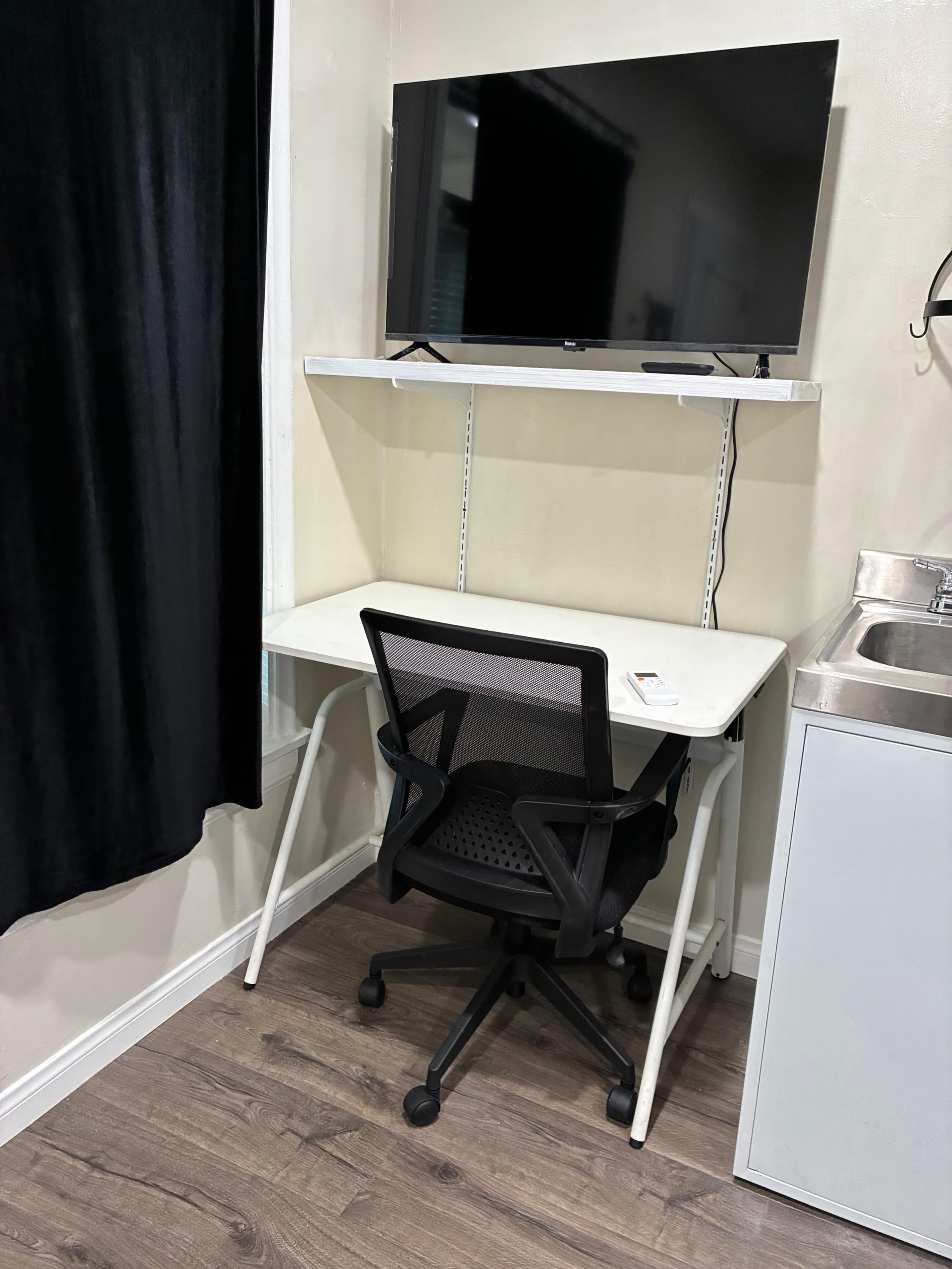 The image shows a small workspace featuring a white desk, a black office chair, a mounted television above a shelf, and a nearby sink.