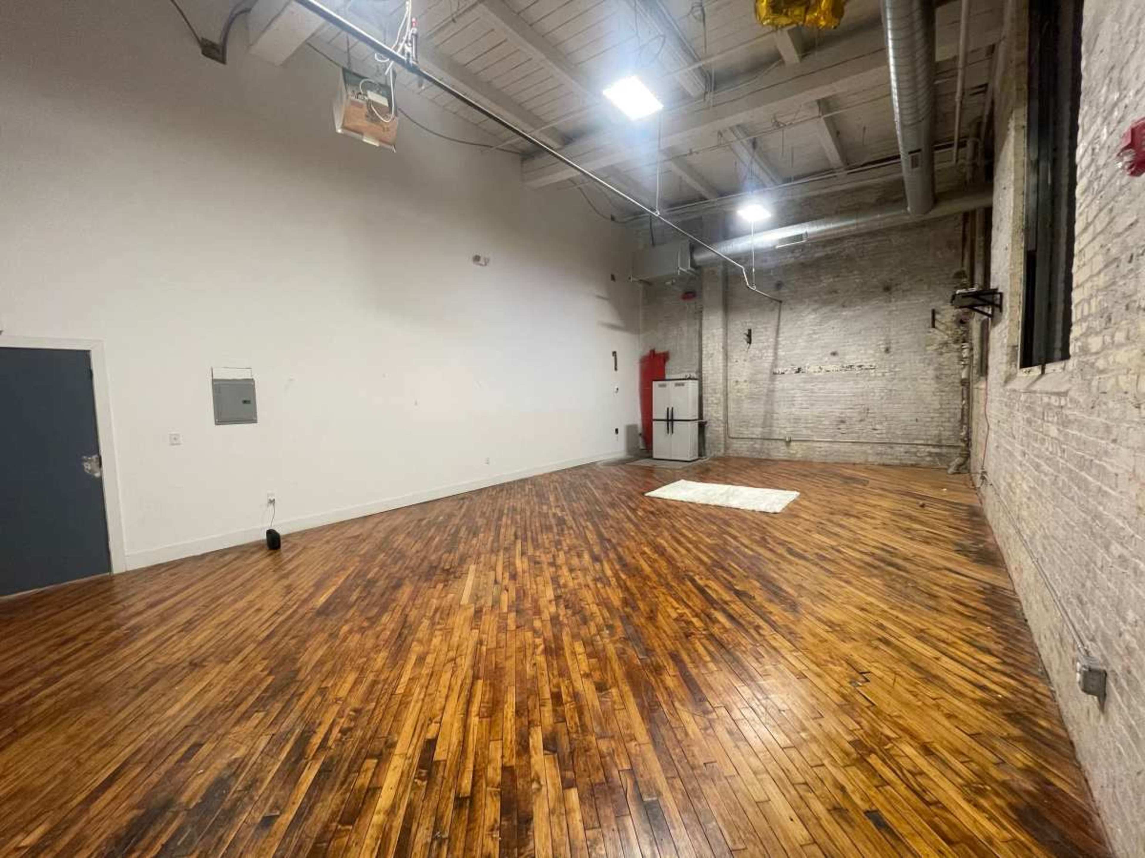 The image shows a spacious, empty room with wooden flooring and exposed brick walls, featuring a single white rug in the center.
