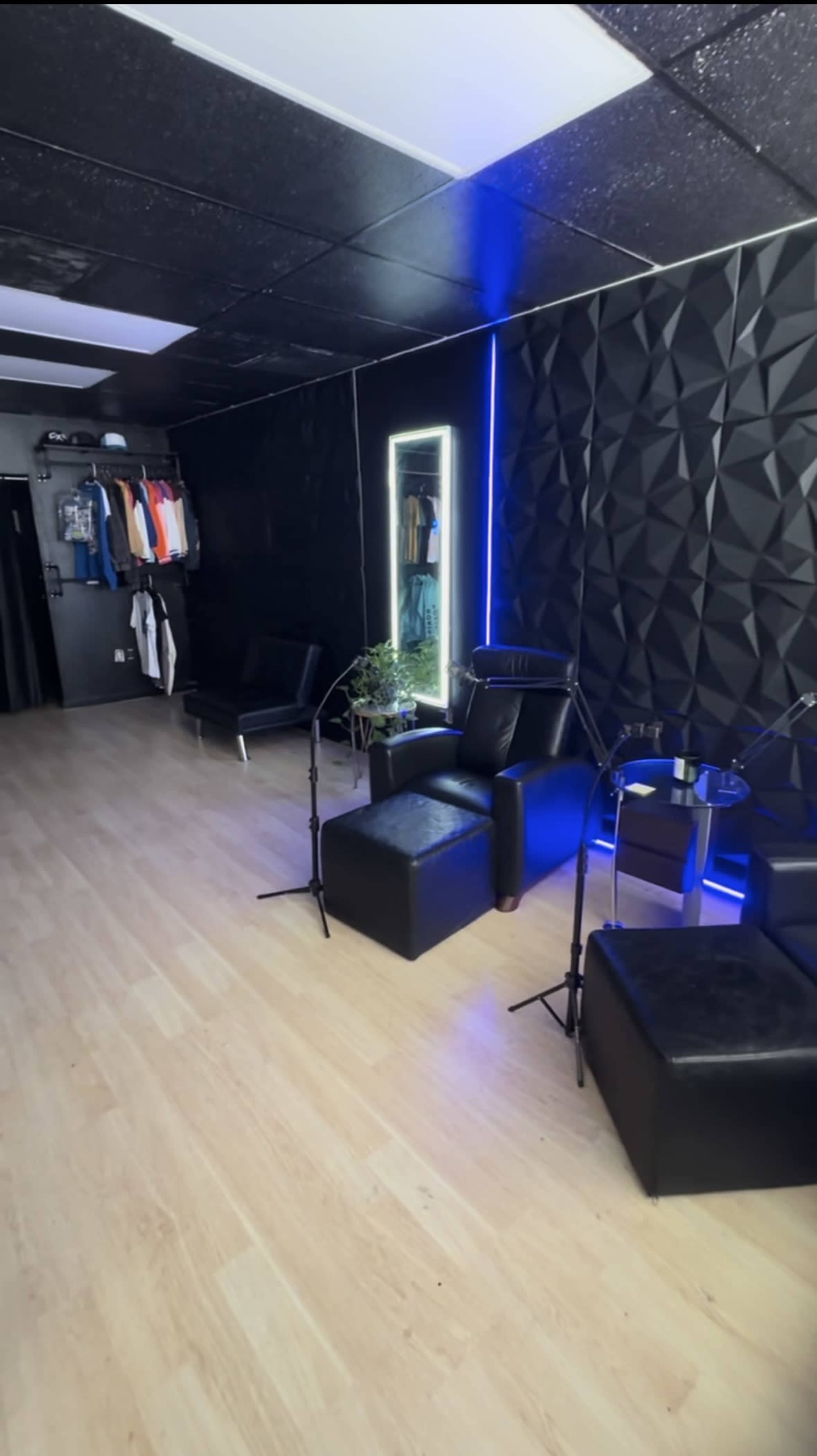 The image shows a modern, minimalist interior of a room with black walls, wooden flooring, and two black chairs positioned near a lit mirror.