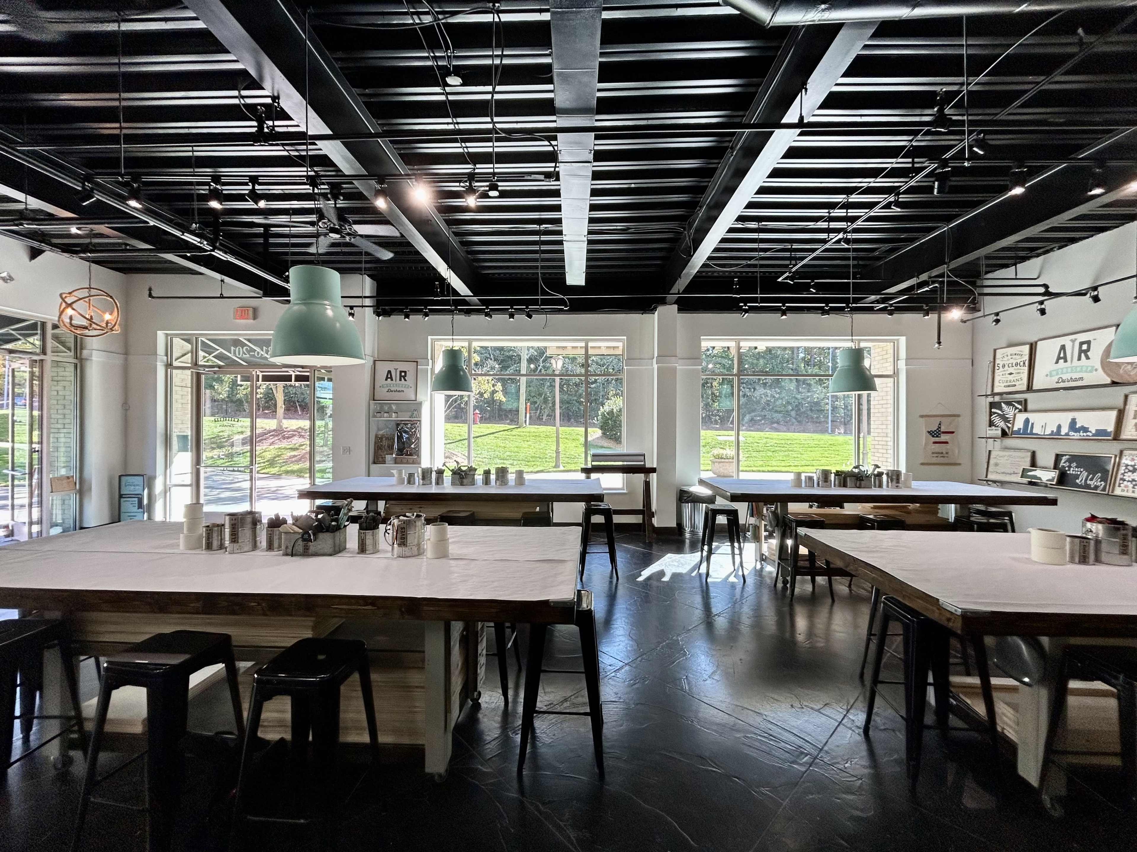 A spacious, modern café with large windows, wooden tables, and metal stools, illuminated by pendant lights.
