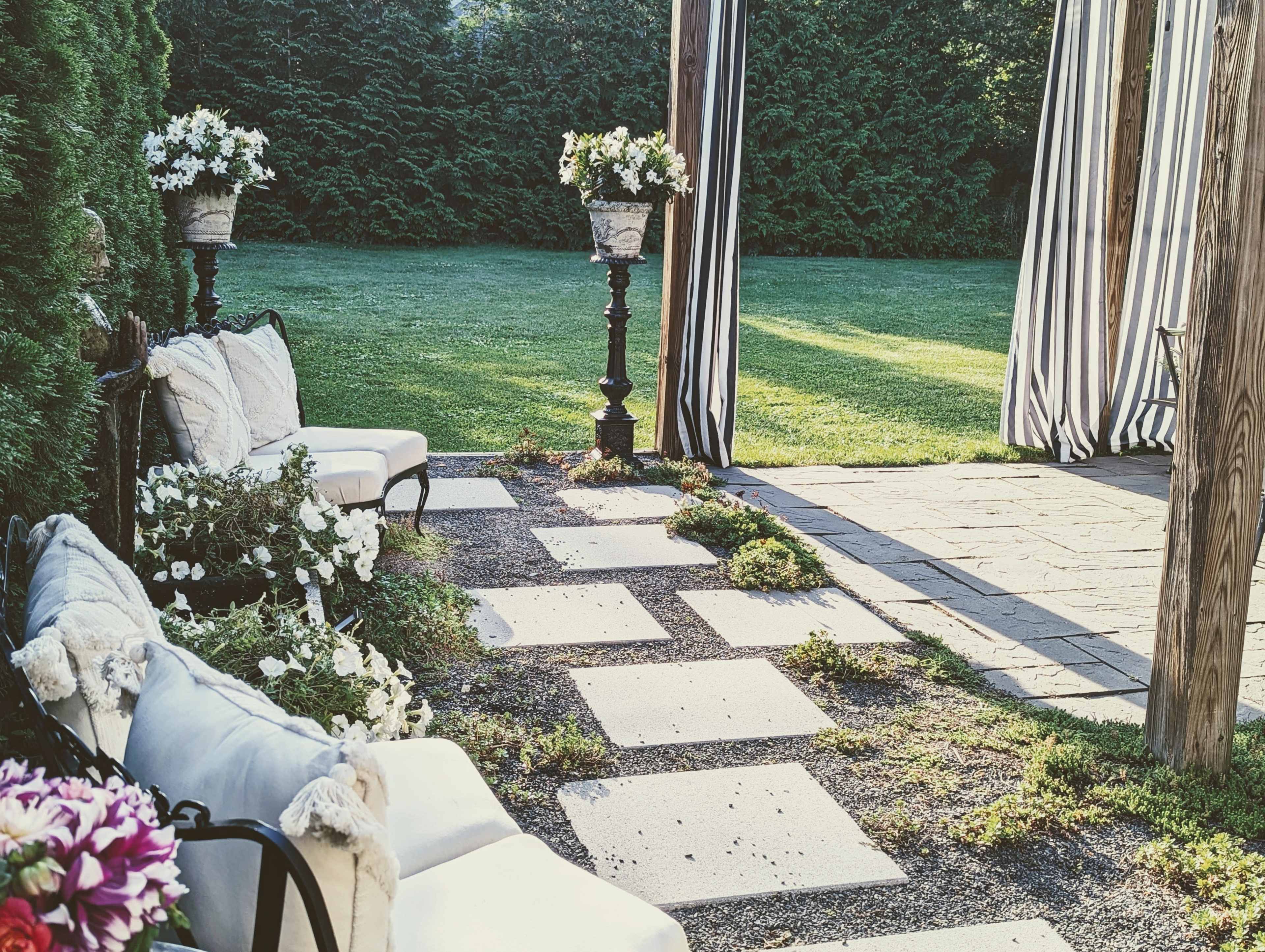A landscaped outdoor space features stone pathways, seating arrangements with pillows, flower planters, and a shaded area defined by striped fabric.