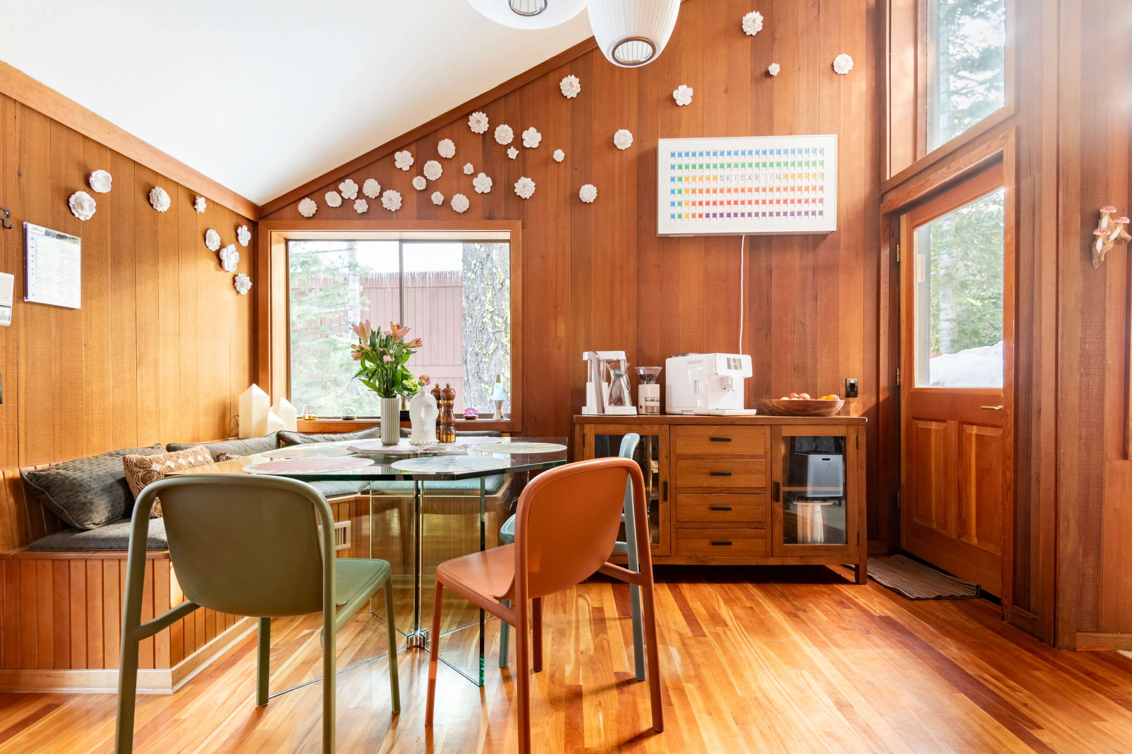 A cozy dining area with wooden walls, a round glass table, and colorful chairs, surrounded by decorative paper flowers and a vibrant wall art piece.