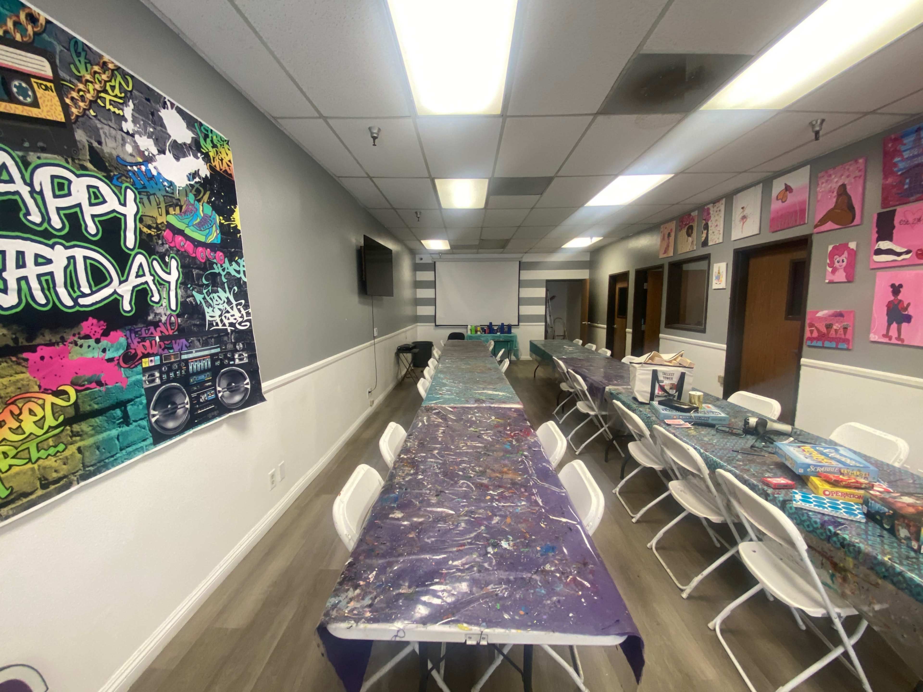 The image shows a neatly arranged event space with several long tables covered in colorful tablecloths, flanked by chairs and decorated walls featuring artwork and a "Happy Birthday" banner.