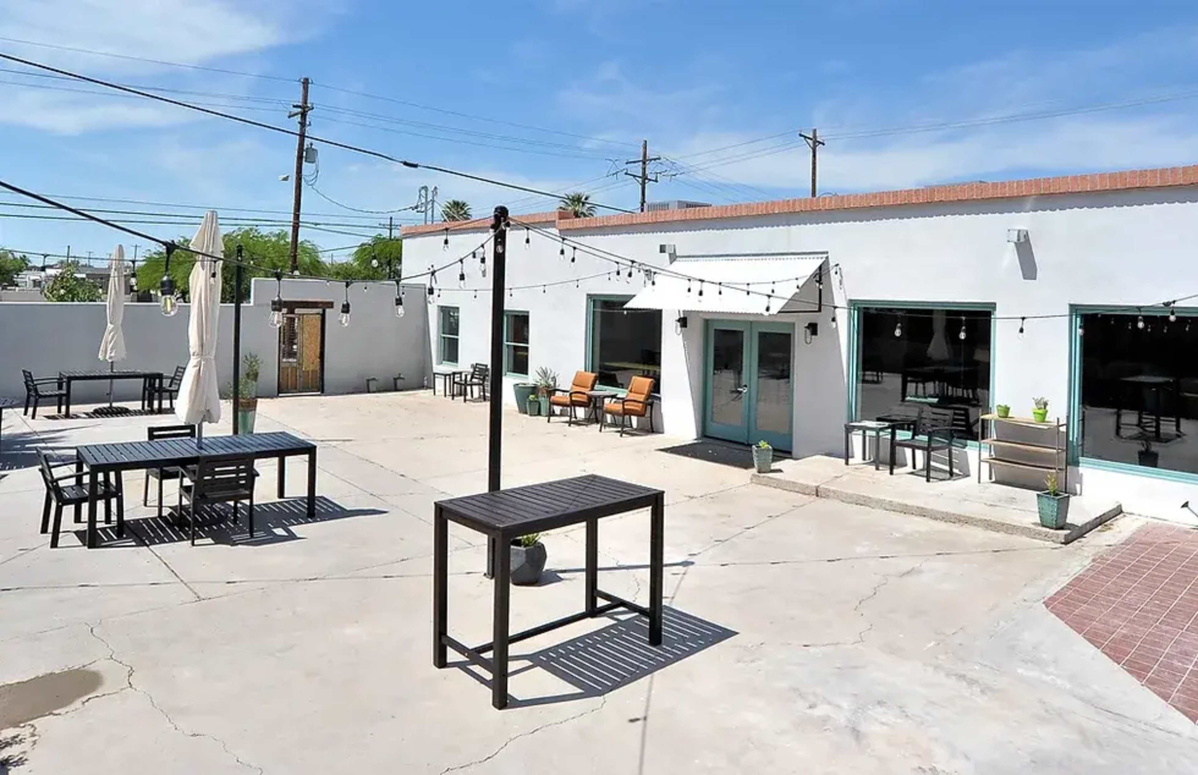 The image shows a spacious outdoor patio with several tables and chairs, string lights overhead, and a building in the background.