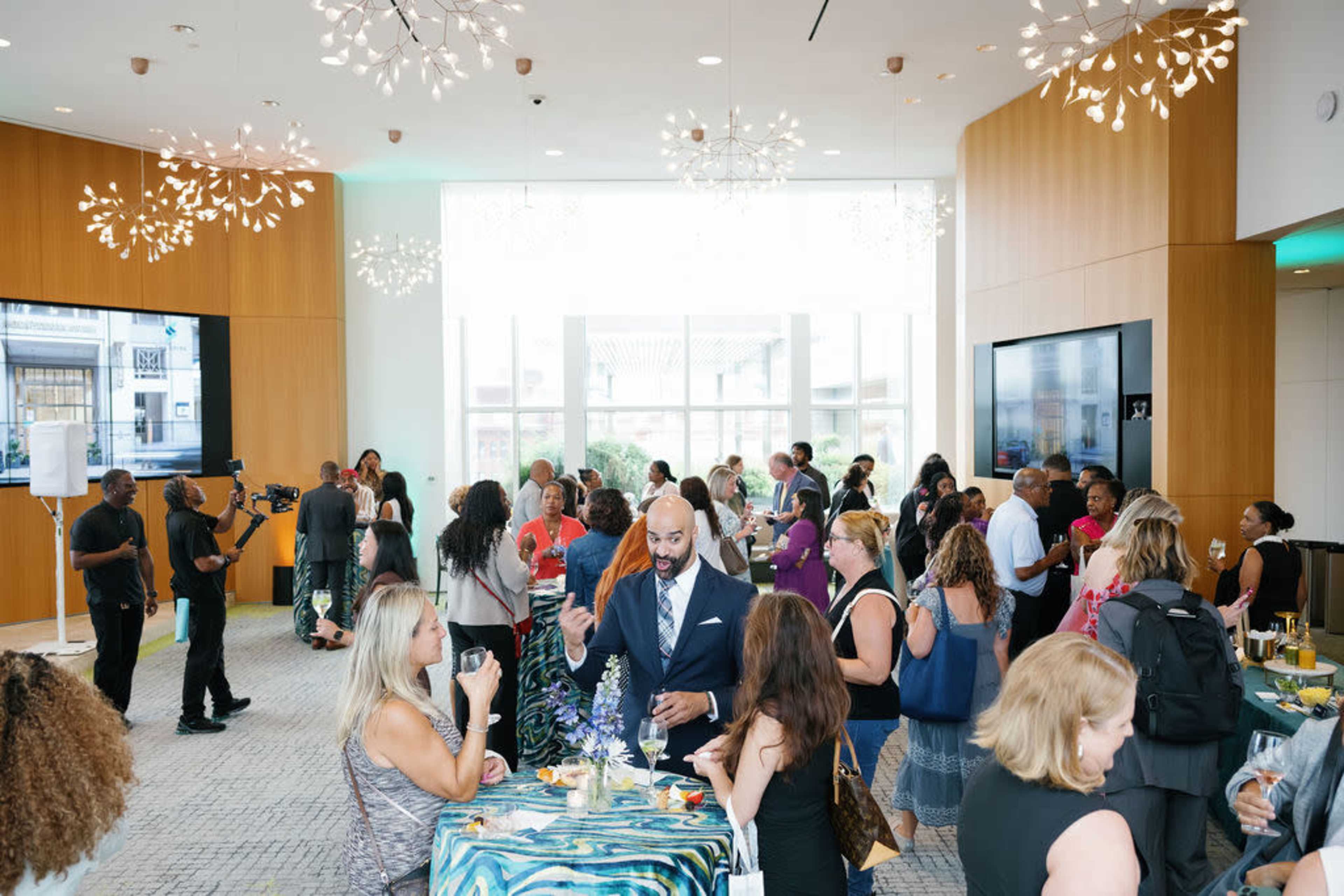 A crowded event space features attendees engaged in conversation and networking, with refreshments available on a table and decorative light fixtures above.