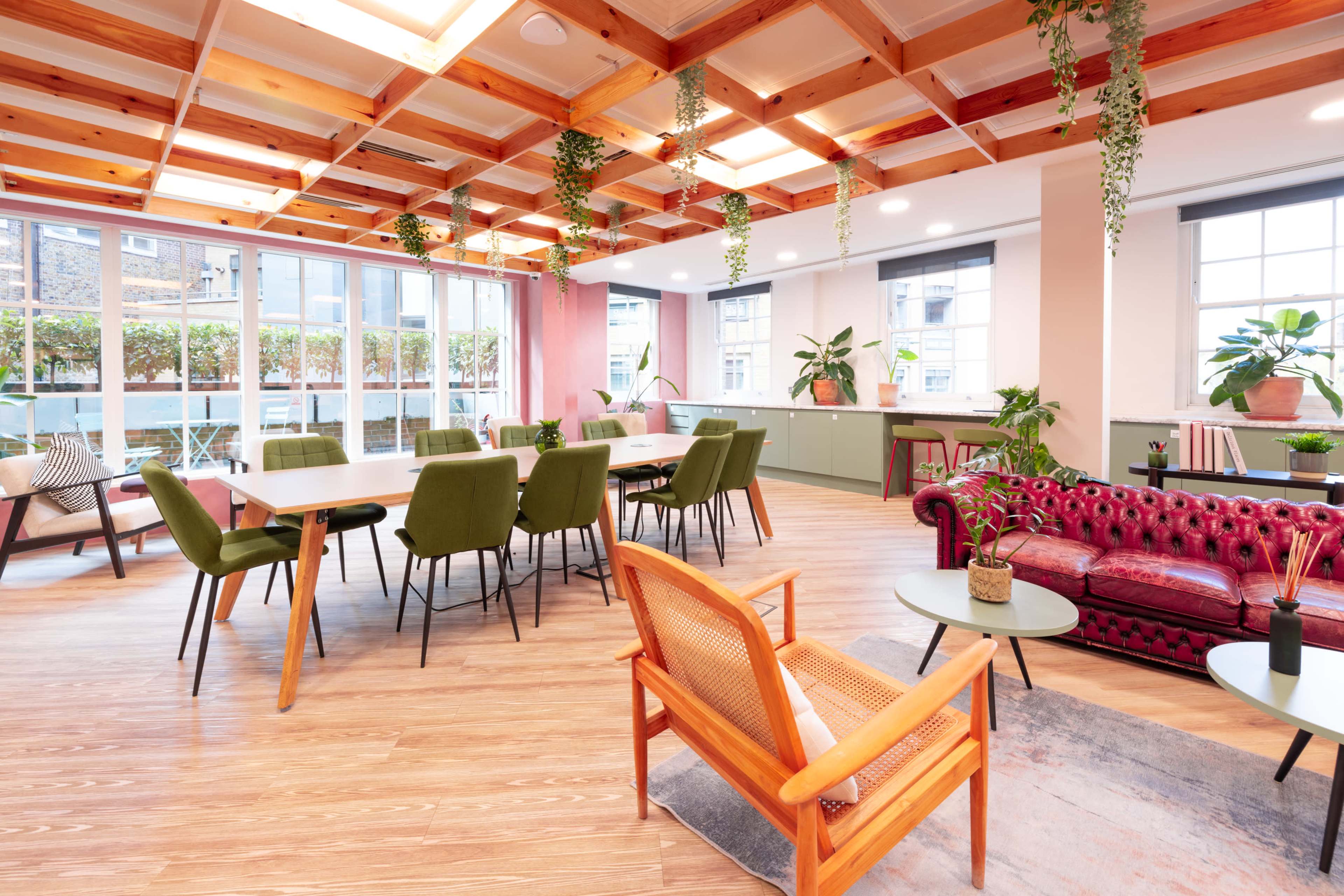 The image shows a modern, open office space featuring a mix of seating options, including a red leather sofa, wooden chairs, and green plants, with large windows providing natural light.
