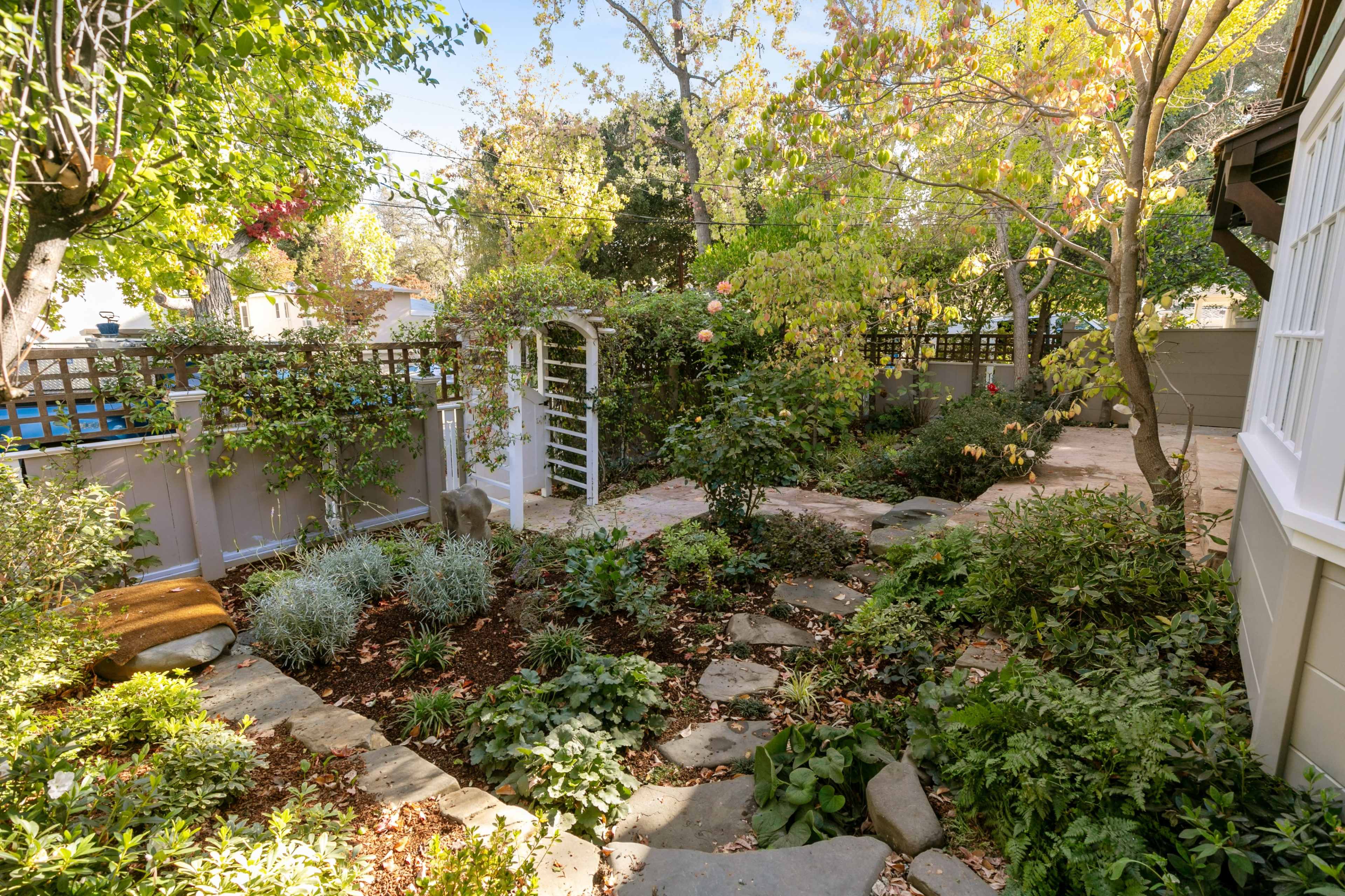 A landscaped backyard featuring a stone pathway, lush greenery, and a white archway surrounded by various plants.